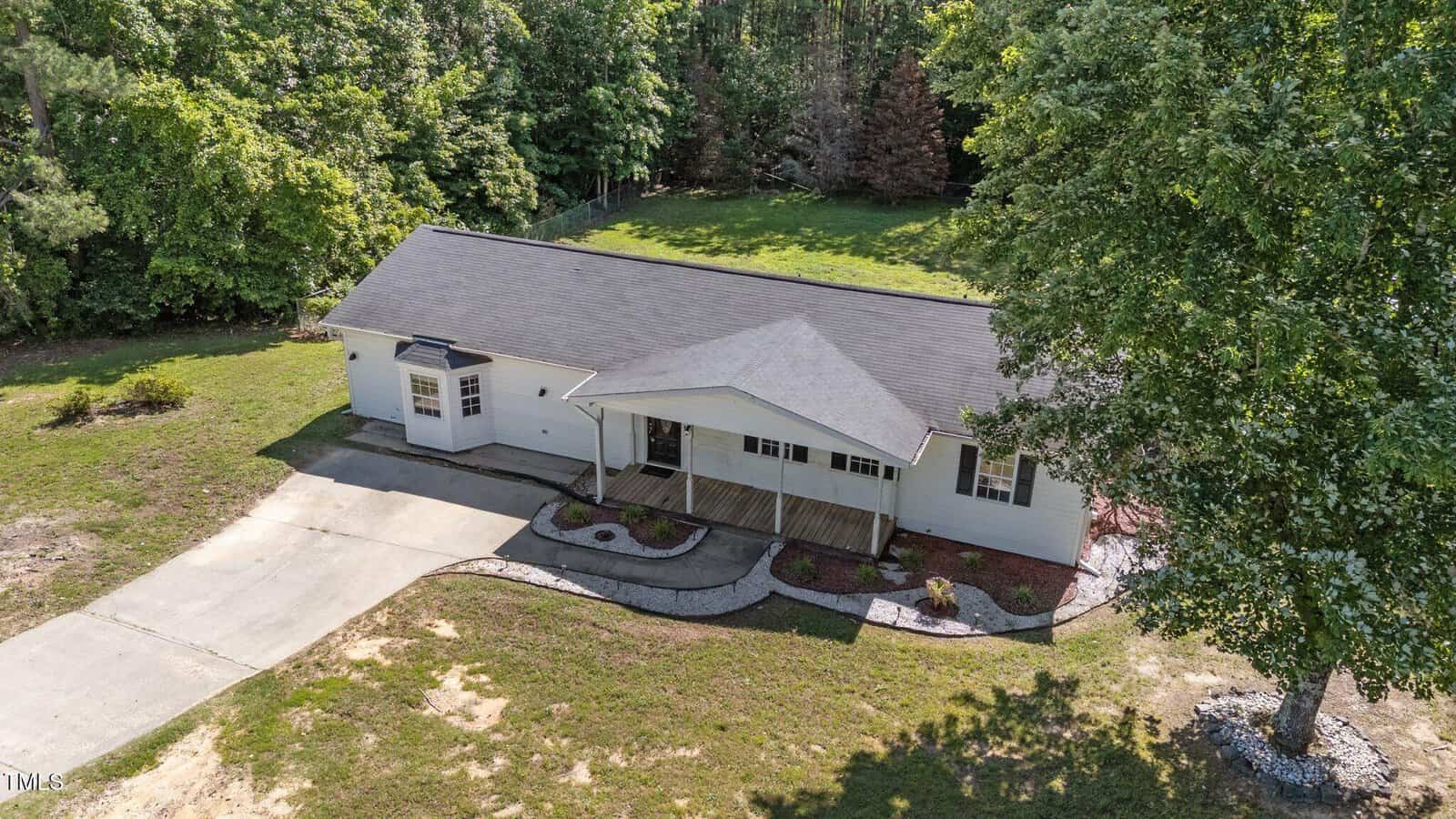 Aerial view of a single-story white house with a covered front porch, attached driveway, and surrounding green lawn, bordered by trees in the background.