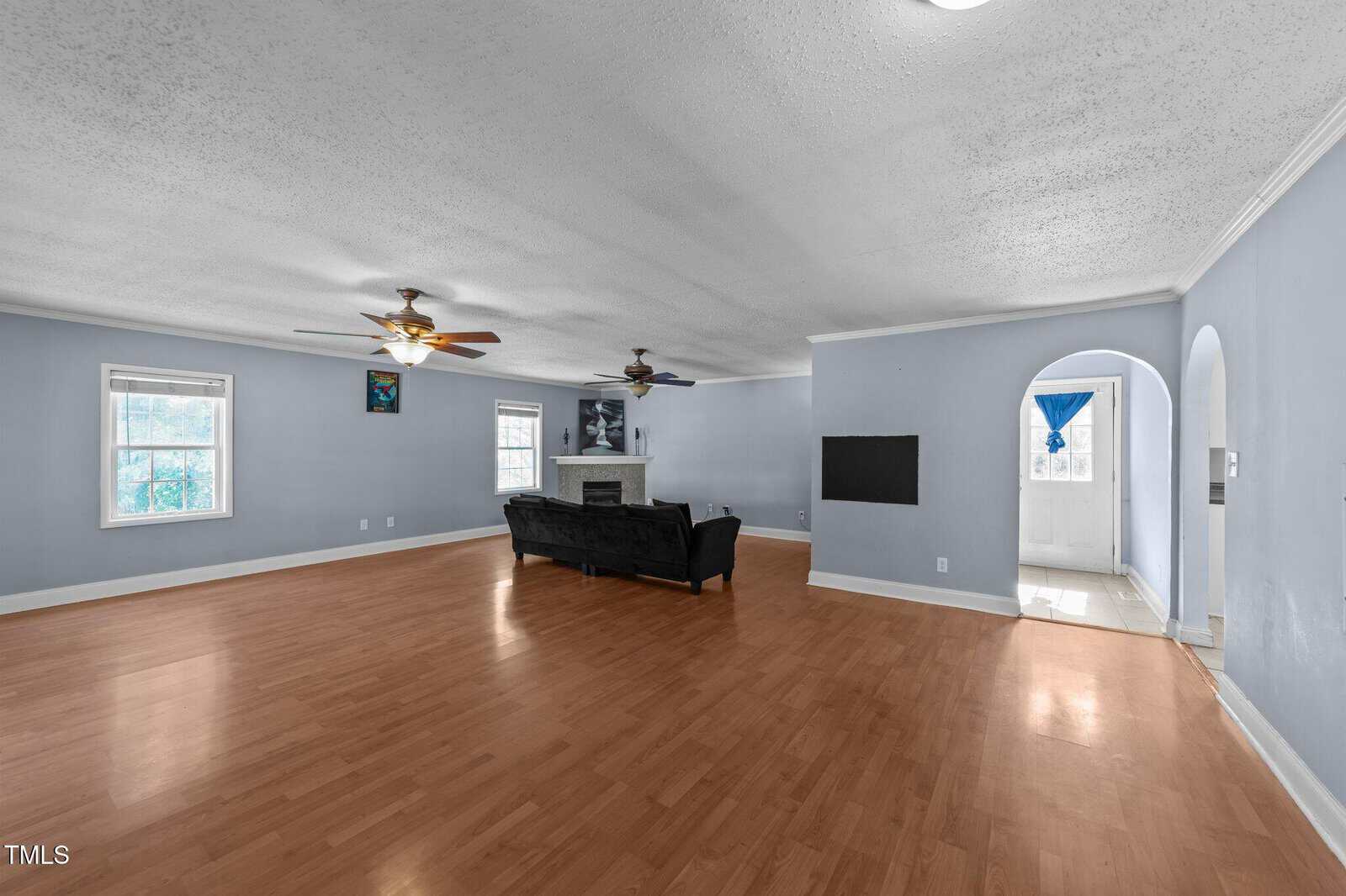 Spacious living room with light wood flooring, pale blue walls, two ceiling fans, and a fireplace. A black couch faces the fireplace. Two windows and an arched doorway lead to a tiled entry area.