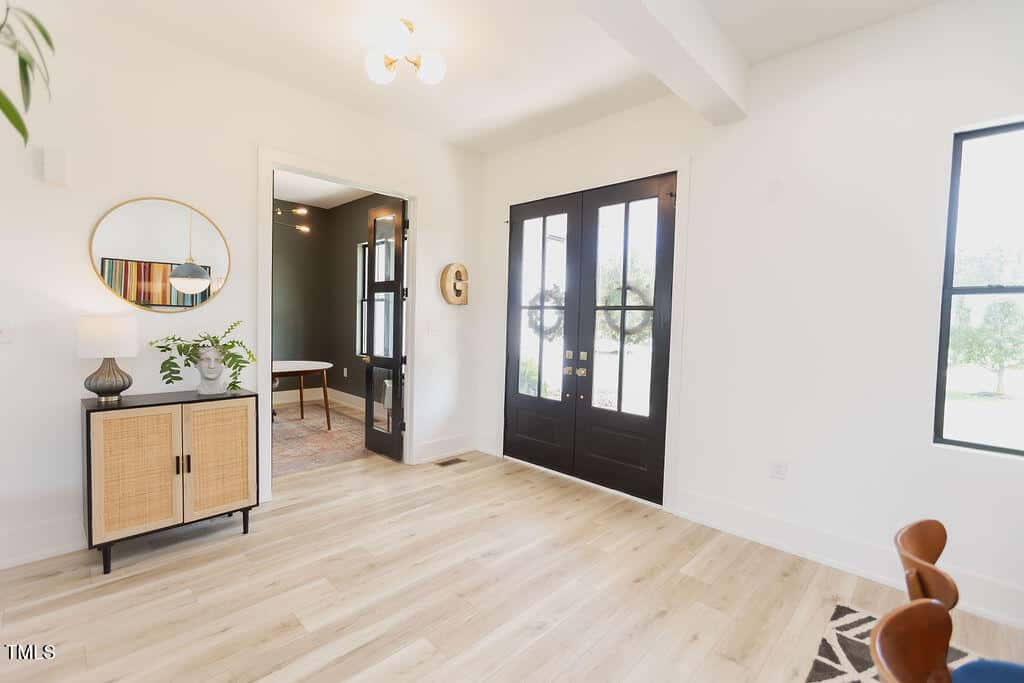Bright, modern entryway with light wood flooring, black double doors, a sideboard with plants and decor, a round mirror above, and windows letting in natural light. A glimpse of an adjacent room is visible.