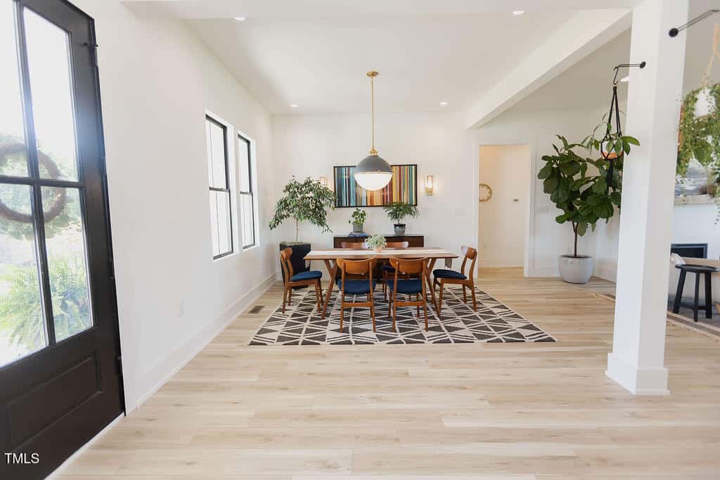 Bright dining room with light wood floors, a geometric rug, a wooden table with six chairs, large windows, potted plants, a hanging pendant light, and modern decor in a spacious, open layout.