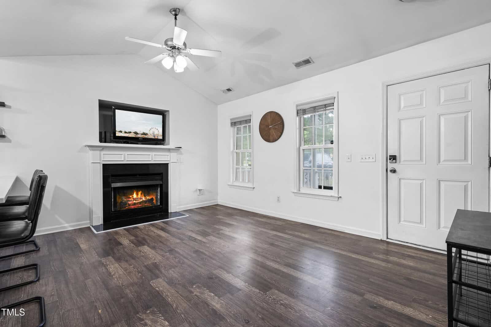 A living room with dark wood floors, a white ceiling fan, a fireplace with a TV above it, two windows with blinds, a round wall clock, a white door, and part of a dining table with chairs visible on the left.