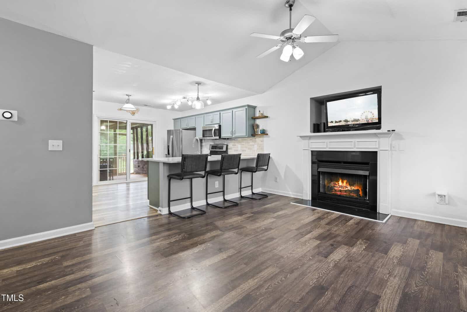 Open-concept living area with dark wood floors, a white fireplace, and a kitchen featuring blue cabinets, stainless steel appliances, a breakfast bar with three stools, and sliding glass doors leading outside.