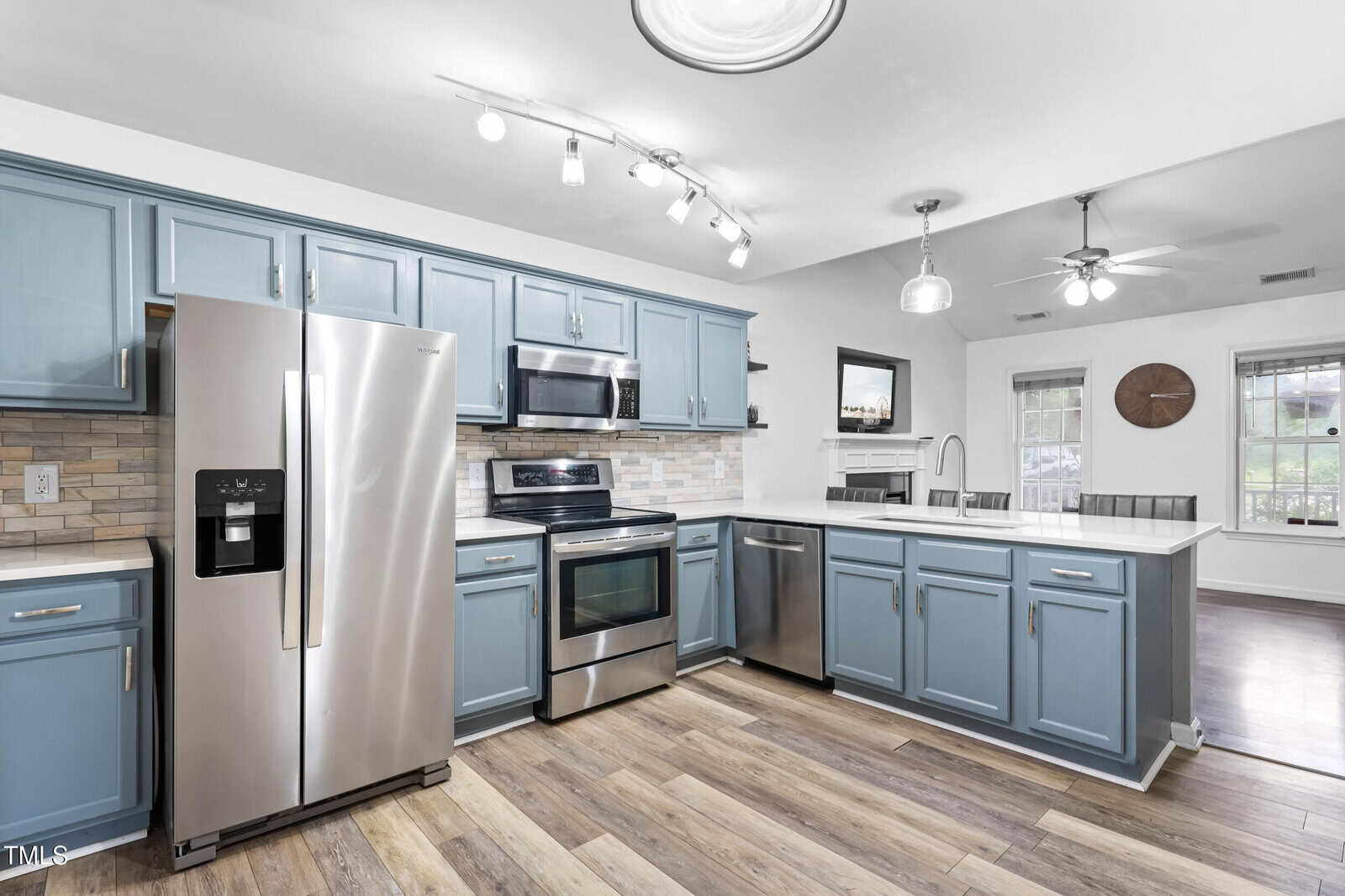 Modern kitchen with stainless steel appliances, blue cabinets, light wood floors, white countertops, and a tile backsplash. Ceiling lights illuminate the space, which opens to a bright living area with windows.
