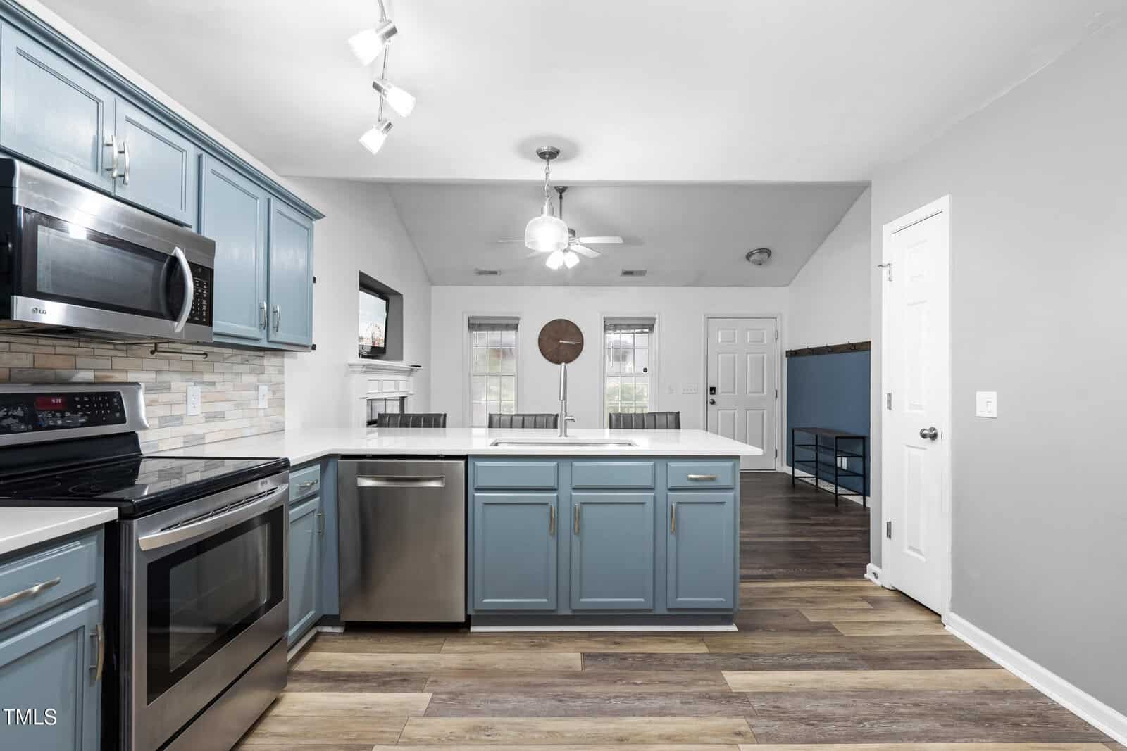 Modern kitchen with blue cabinets, stainless steel appliances, white countertops, tile backsplash, wood-style flooring, and an open layout leading to a living area with large windows and a ceiling fan.