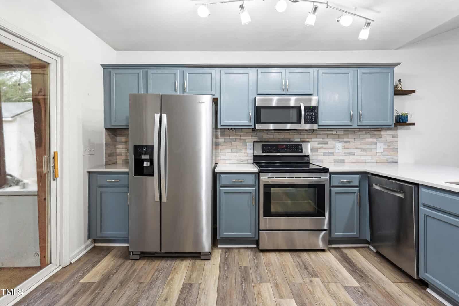 Modern kitchen with stainless steel appliances, blue cabinets, a tile backsplash, under-cabinet lighting, and wood-look flooring. Refrigerator, microwave, oven, and dishwasher are visible. Countertops are white.