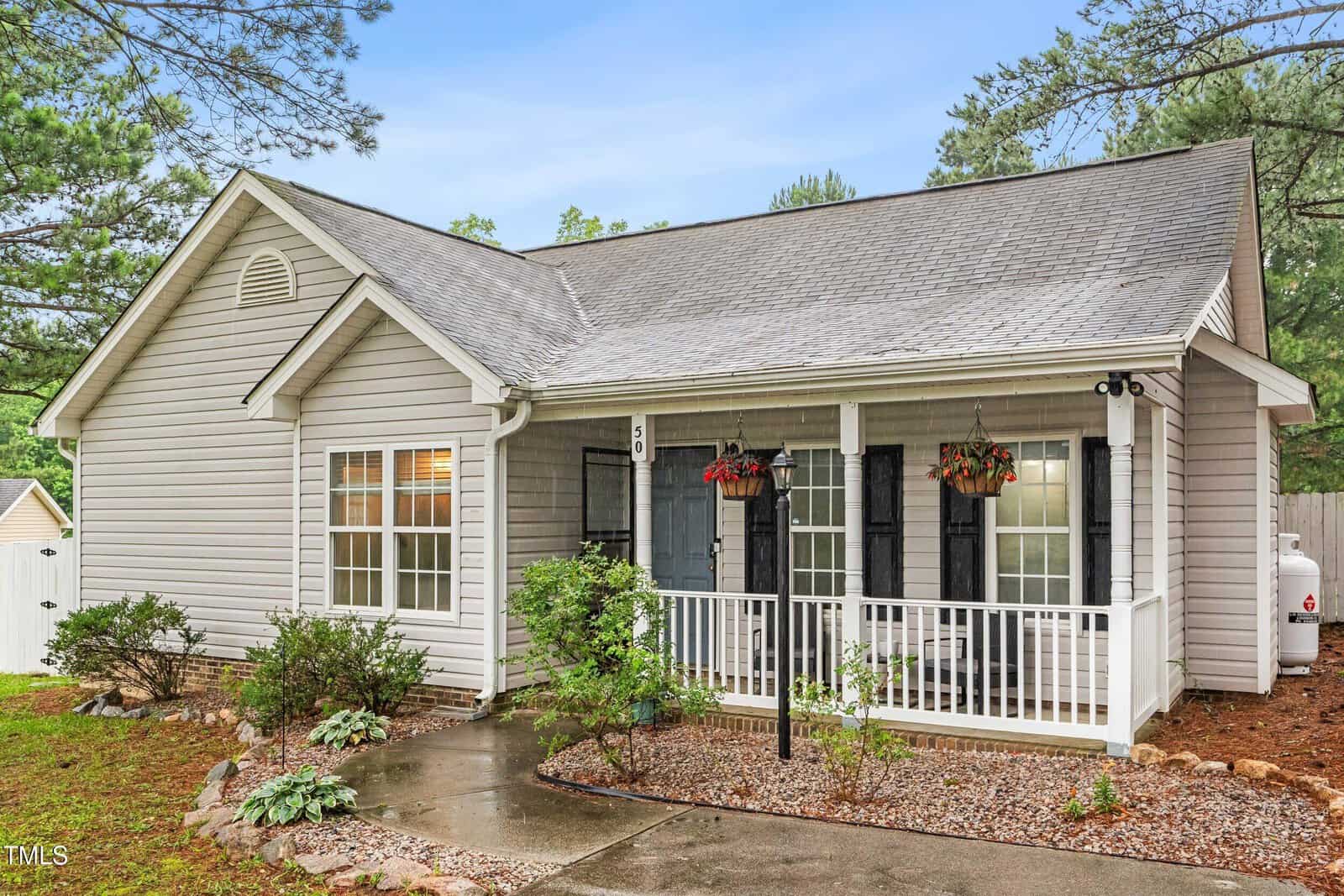 A single-story gray house with white trim, a covered front porch with hanging plants, black shutters, and a small garden along a walkway, surrounded by trees.