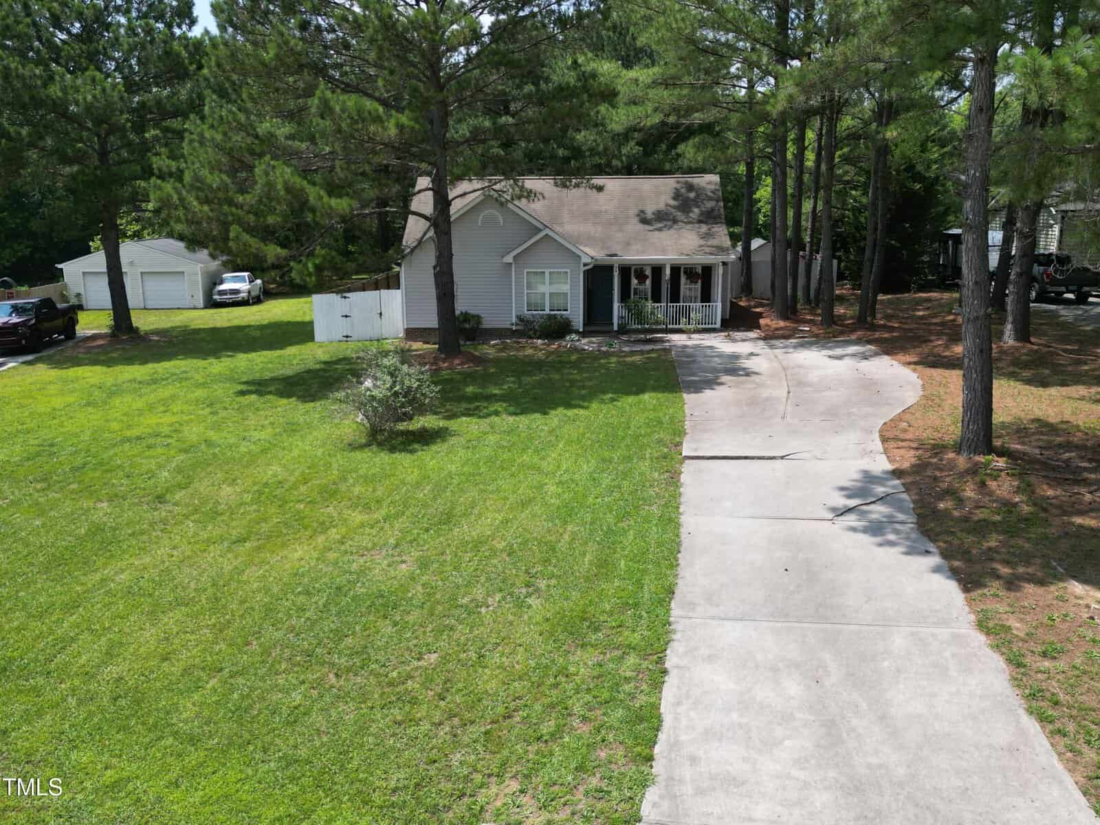 A small gray house with white trim sits among tall pine trees, featuring a front porch and curved driveway leading up to it. The yard is spacious with green grass and a white shed in the back.