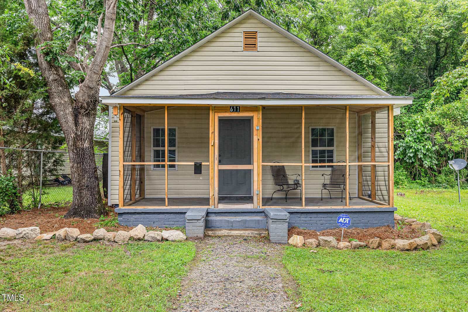 A small beige house at 611 Rowland Street, Henderson, features a screened front porch with two chairs and a dark front door. A stone path leads to the steps, an ADT security sign is by the porch, and trees and greenery surround the home.