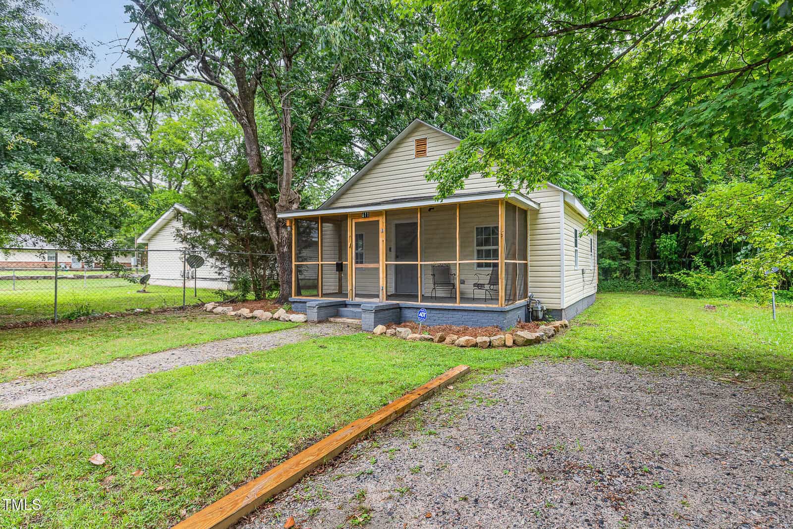 Single-story house with a screened front porch, beige siding, and dark trim, surrounded by a grassy yard with a stone border and mature trees. A gravel path leads to the porch, and a chain-link fence is visible on the left.