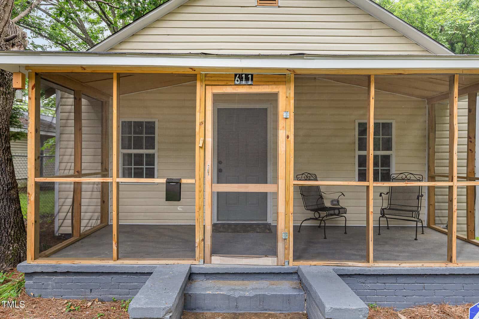 A beige house with a screened-in front porch, two metal chairs, a small table, and steps leading up to a gray front door. The porch is supported by wooden frames, and there are trees nearby.