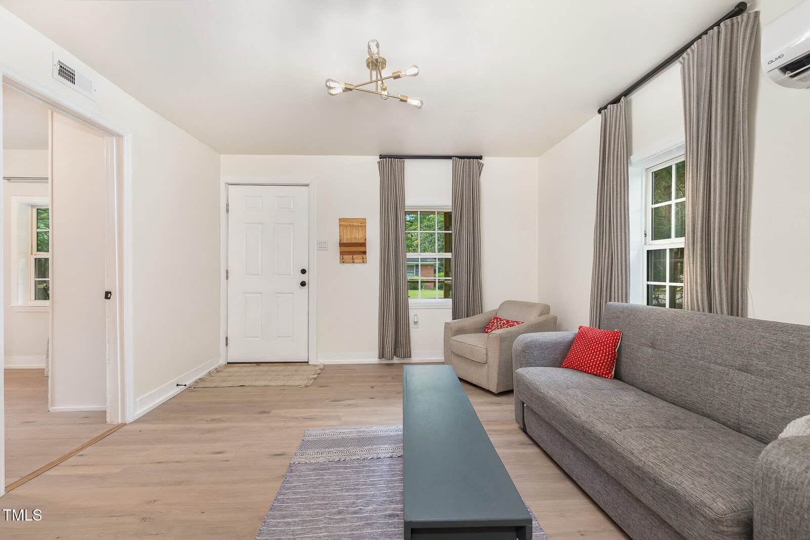 A living room with light wood flooring, a gray sofa with a red cushion, a beige armchair, a rectangular coffee table, two windows with gray curtains, and a white front door. A modern ceiling light fixture is overhead.