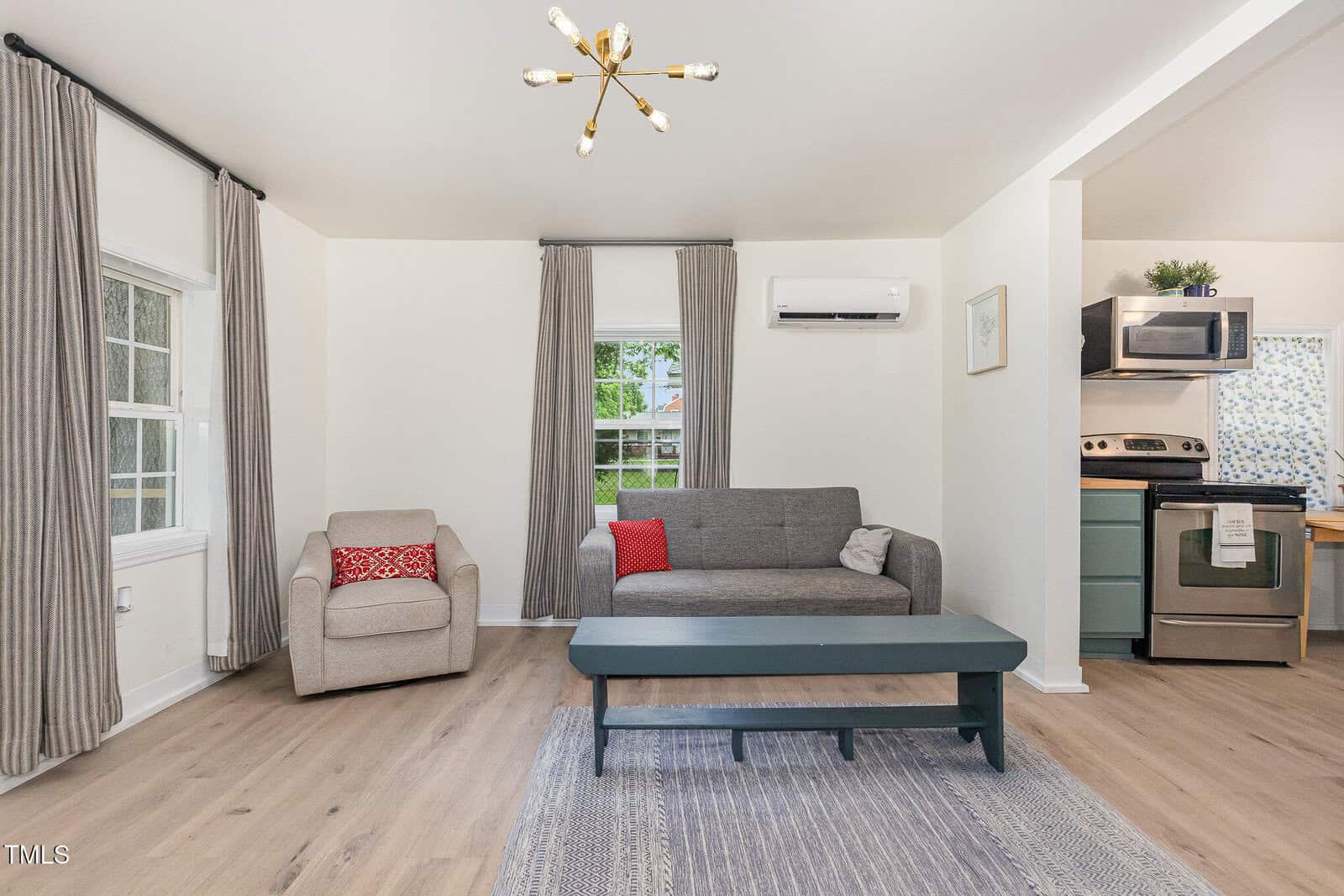A living room with light wood flooring, a gray sofa, a beige armchair, a dark rectangular coffee table, and a rug. There are two windows with gray curtains, an air conditioner, and an open view of a modern kitchen.