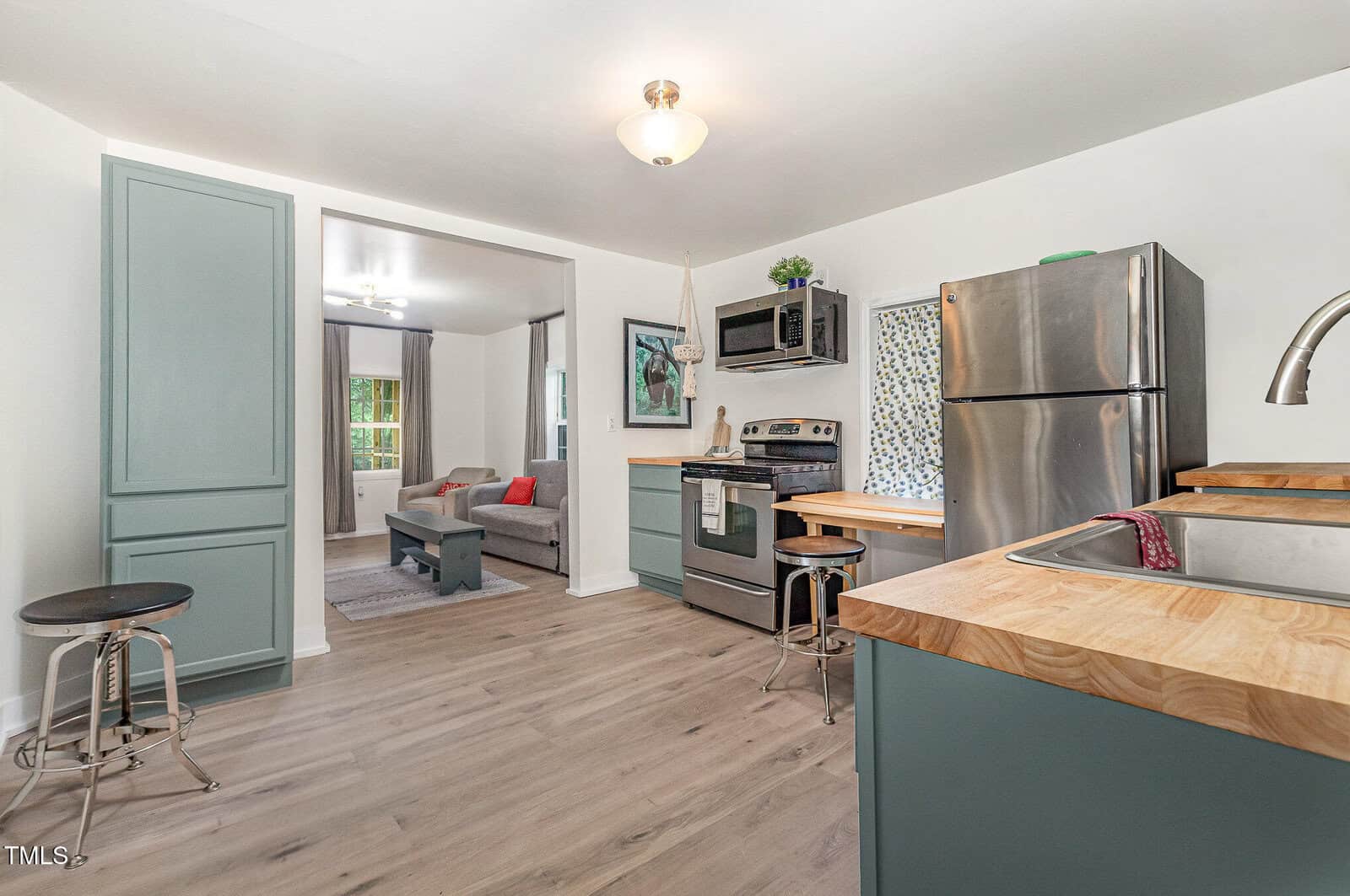 Modern kitchen with green cabinets, stainless steel appliances, wood countertops, and light wood flooring. An open doorway leads to a living room with a gray couch and window. Natural light brightens the clean, airy space.