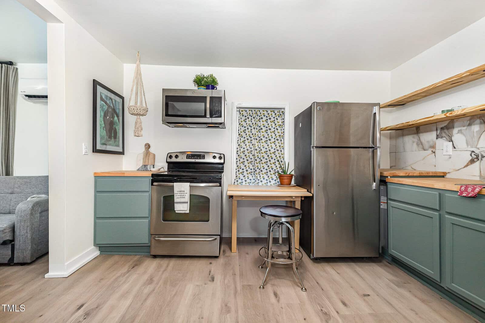 Modern kitchen with stainless steel appliances, green cabinets, wooden countertops, open shelves, a potted plant, and a patterned curtain on a small window. A gray stool sits in front of a wooden table.
