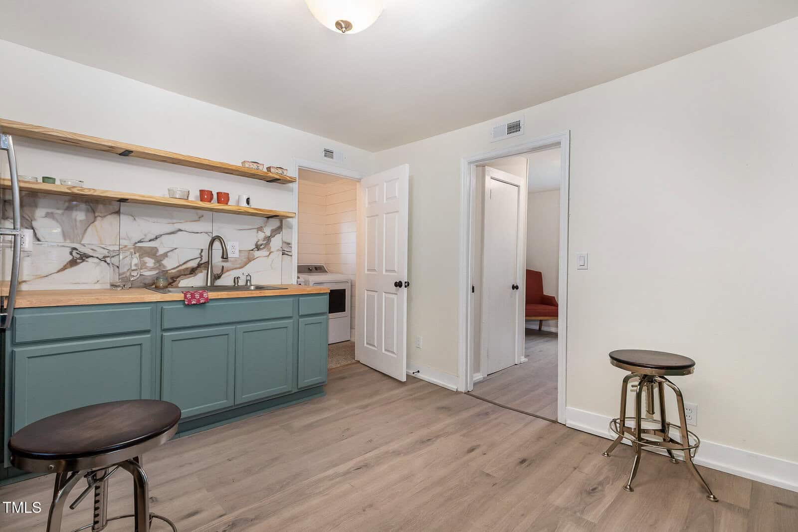 Small kitchen with green cabinets, marble backsplash, open wooden shelves with dishes, two metal stools, and a view into adjoining rooms with white walls and light wood flooring.