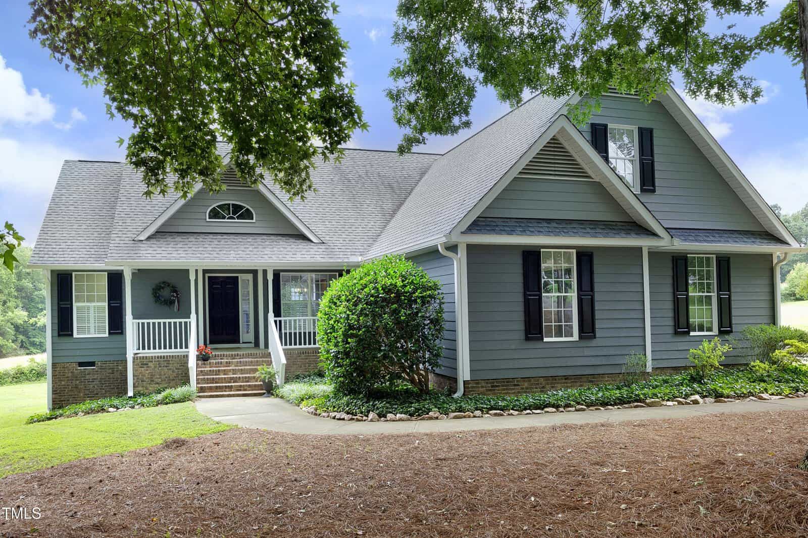 A gray two-story house with white trim and black shutters at 3684 Ben Mil Court, Franklinton, features a covered front porch with white railings and neatly landscaped shrubs along a curved walkway, set against a backdrop of trees and grass.