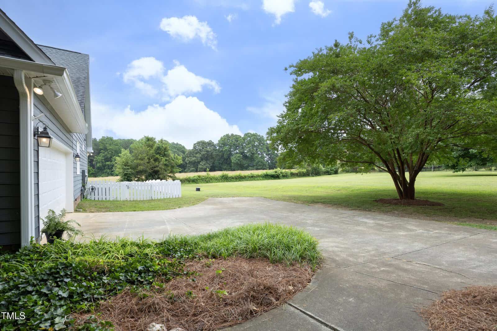 A concrete driveway curves beside a house, with a large tree and green lawn on the right. A white picket fence and landscaped area with mulch and shrubs are in the foreground. Blue sky with scattered clouds overhead.
