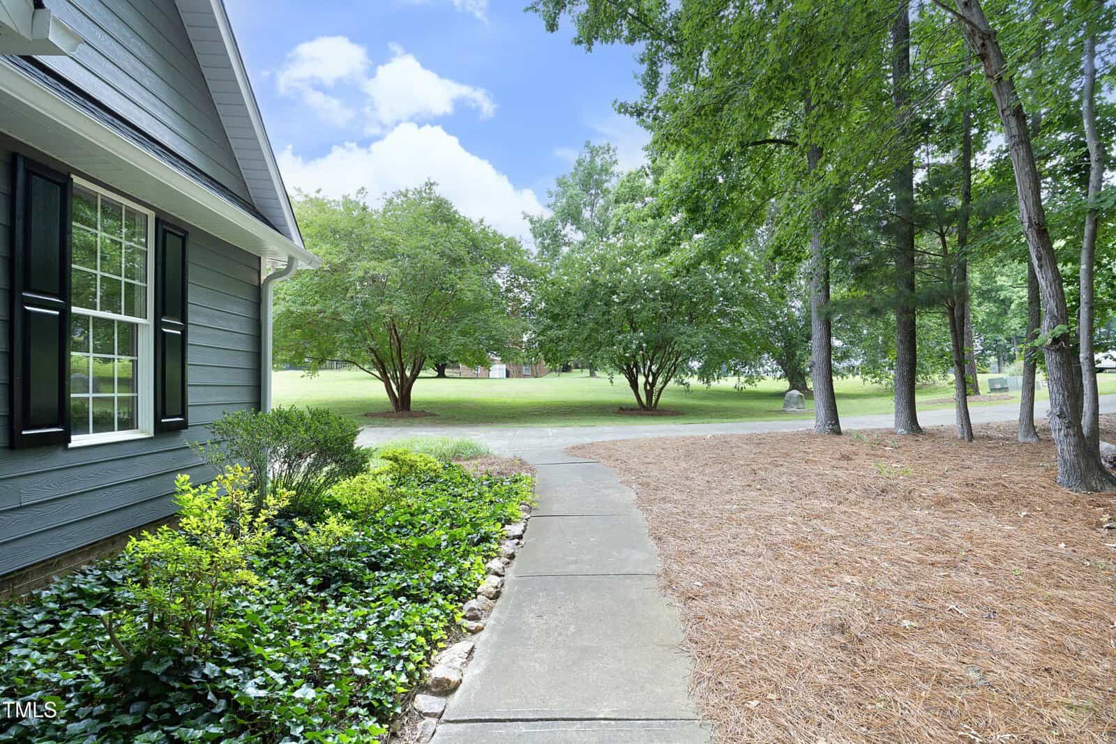 A concrete walkway curves past green shrubs and pine straw mulch beside a blue house, leading toward a grassy yard with trees under a partly cloudy sky.