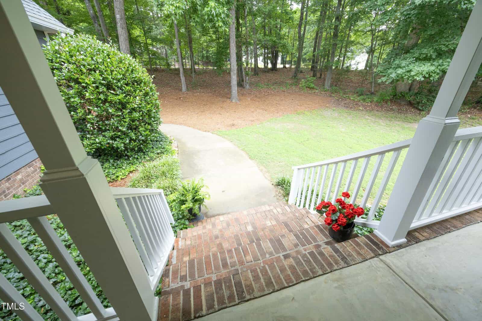 View from a porch with white railings and brick steps leading to a curved sidewalk, surrounded by grass, trees, and a potted plant with red flowers.