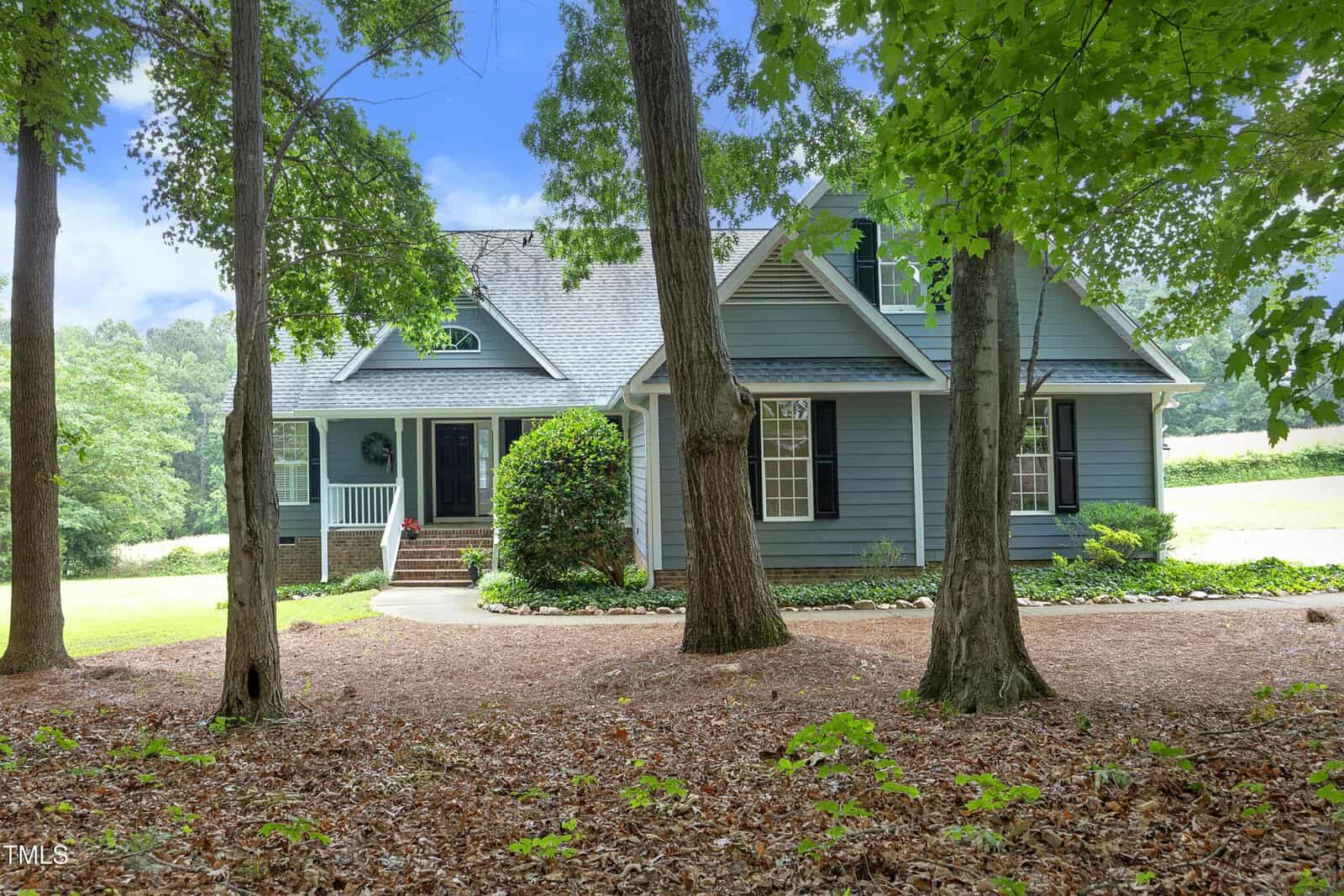 A two-story blue house with white trim and black shutters sits behind several tall trees, with a front porch, brick steps, and a landscaped yard on a sunny day.