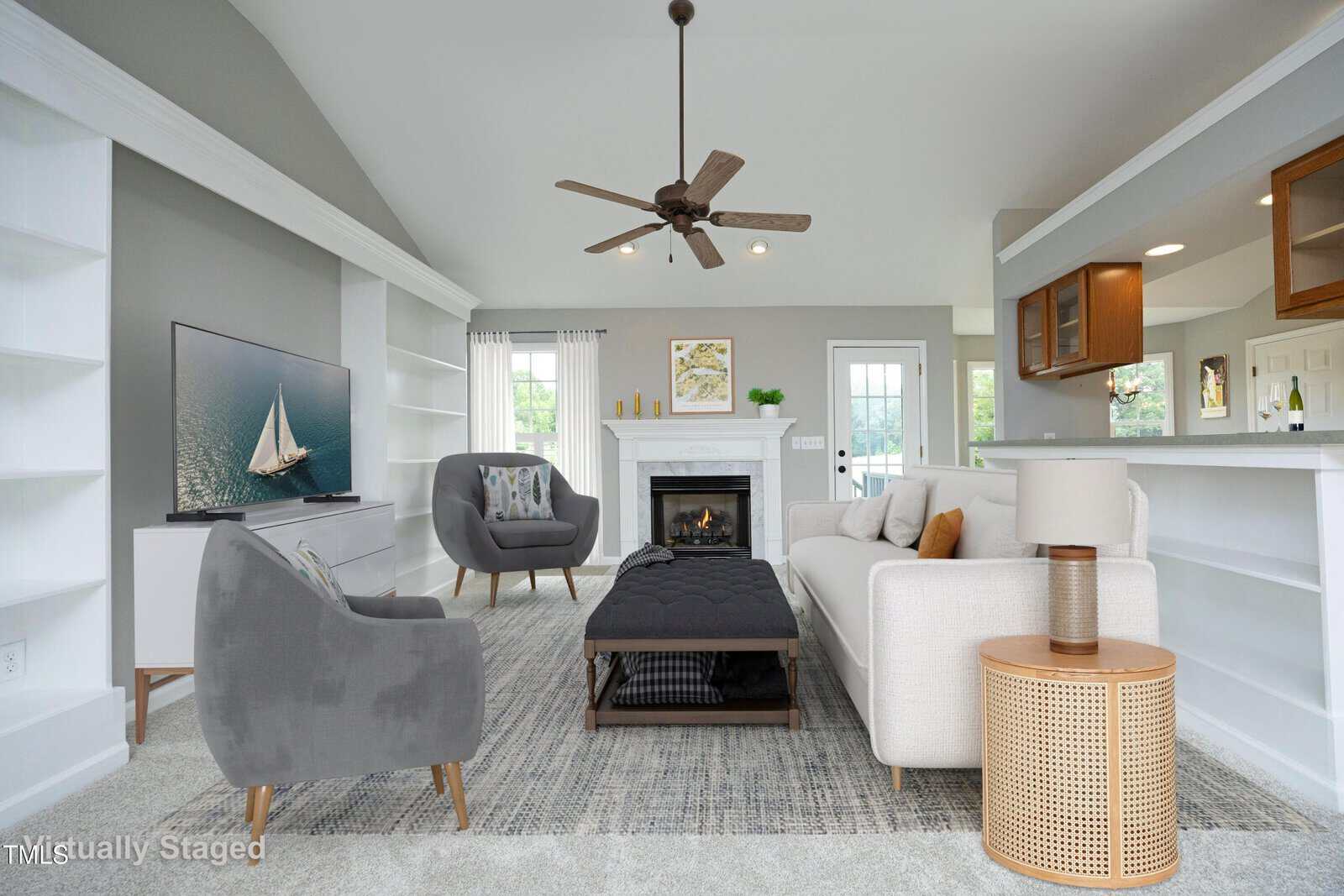 A bright living room features gray and beige furniture, a ceiling fan, a fireplace, a TV with a sailboat image, built-in shelves, and a kitchen pass-through window. The room is virtually staged.