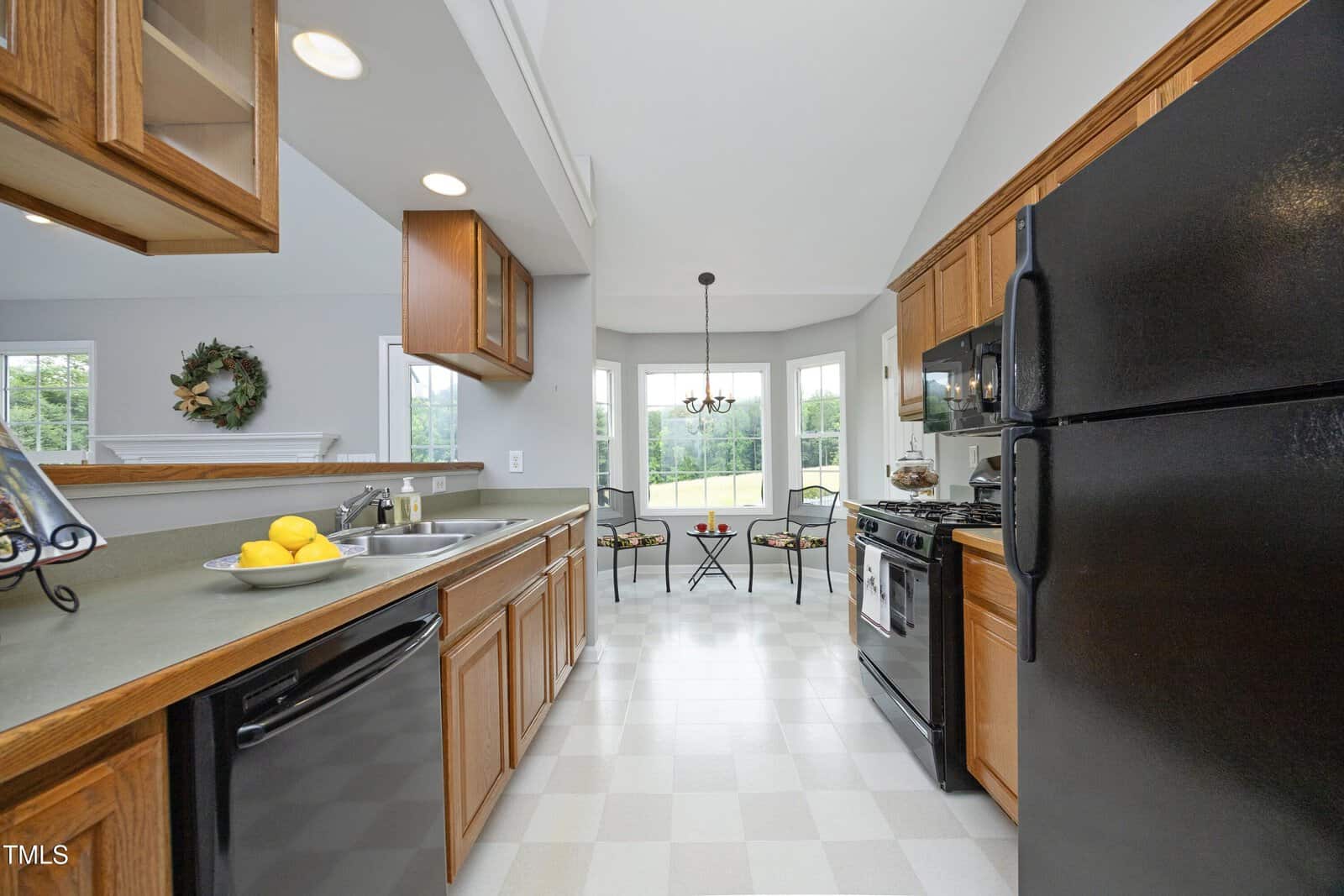 A kitchen with wood cabinets, black appliances, and a gray countertop. A bowl of lemons sits by the sink. In the background, a small dining area with two chairs and a table is positioned by large windows.