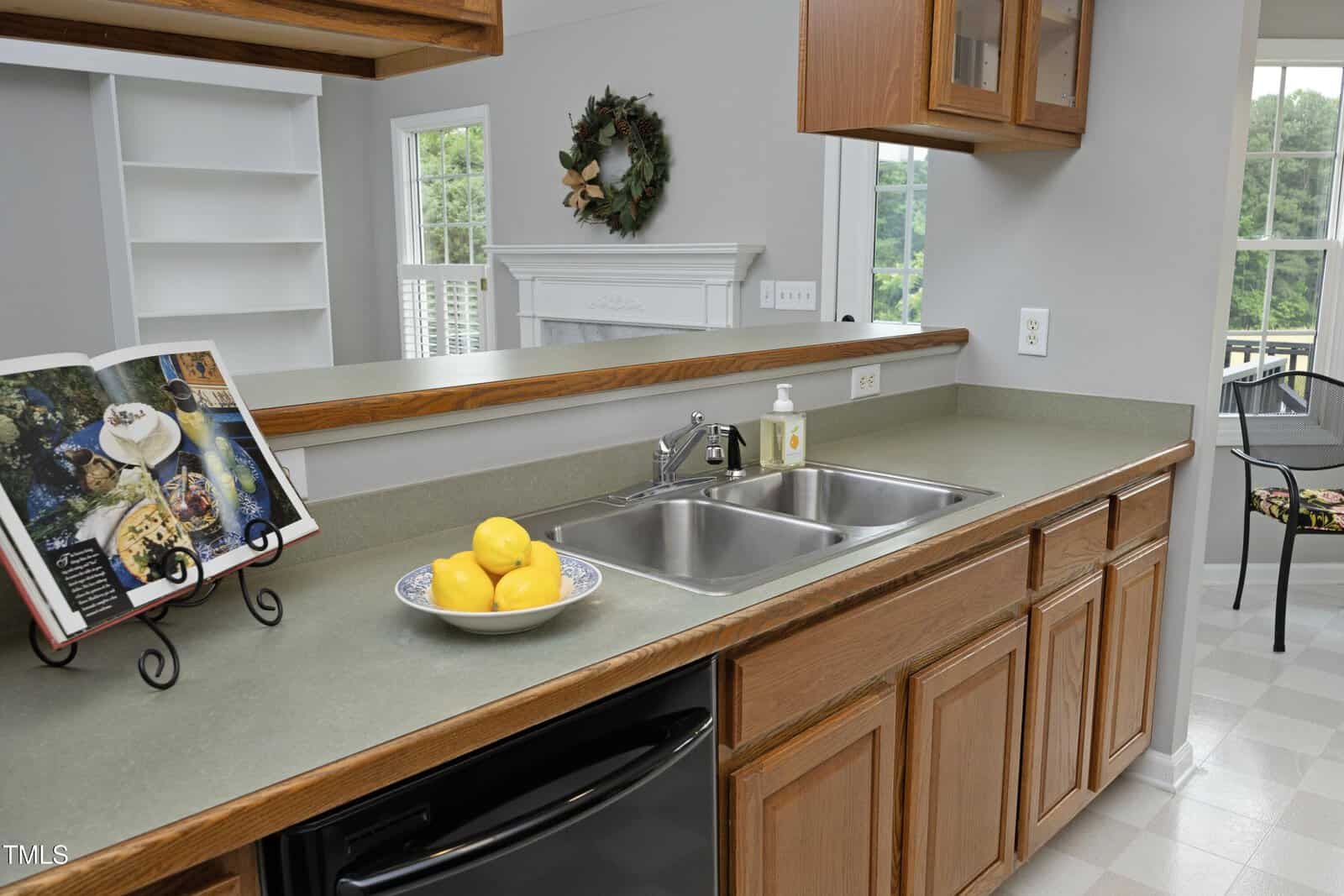 A kitchen countertop with a double sink, a bowl of lemons, a bottle of soap, and an open cookbook on a stand. Wooden cabinets are above and below the counter, and a window and wreath are visible in the background.