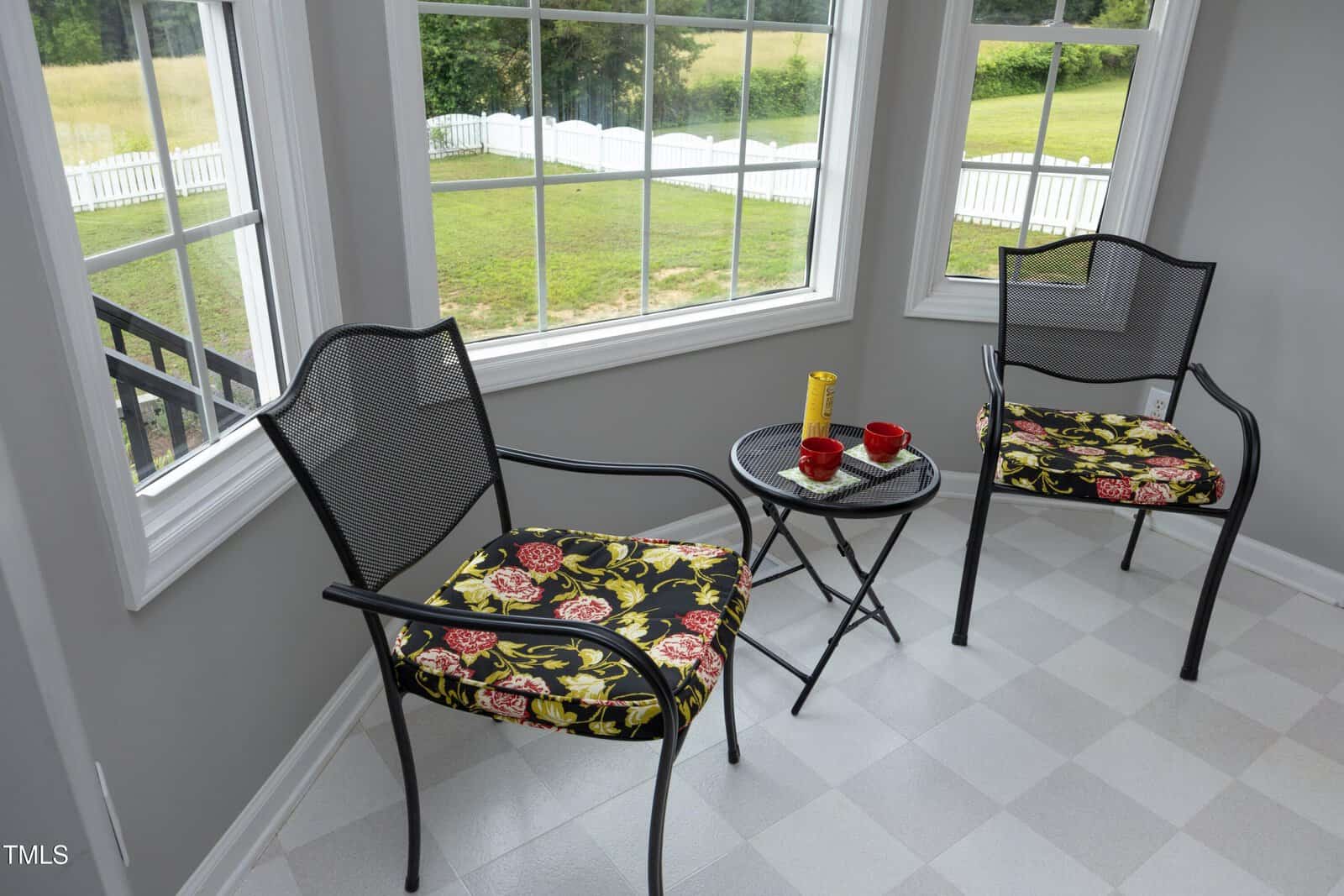 Two black metal chairs with floral cushions and a small round table with two red cups and a yellow thermos are set in a sunroom with large windows overlooking a grassy yard and white fence.