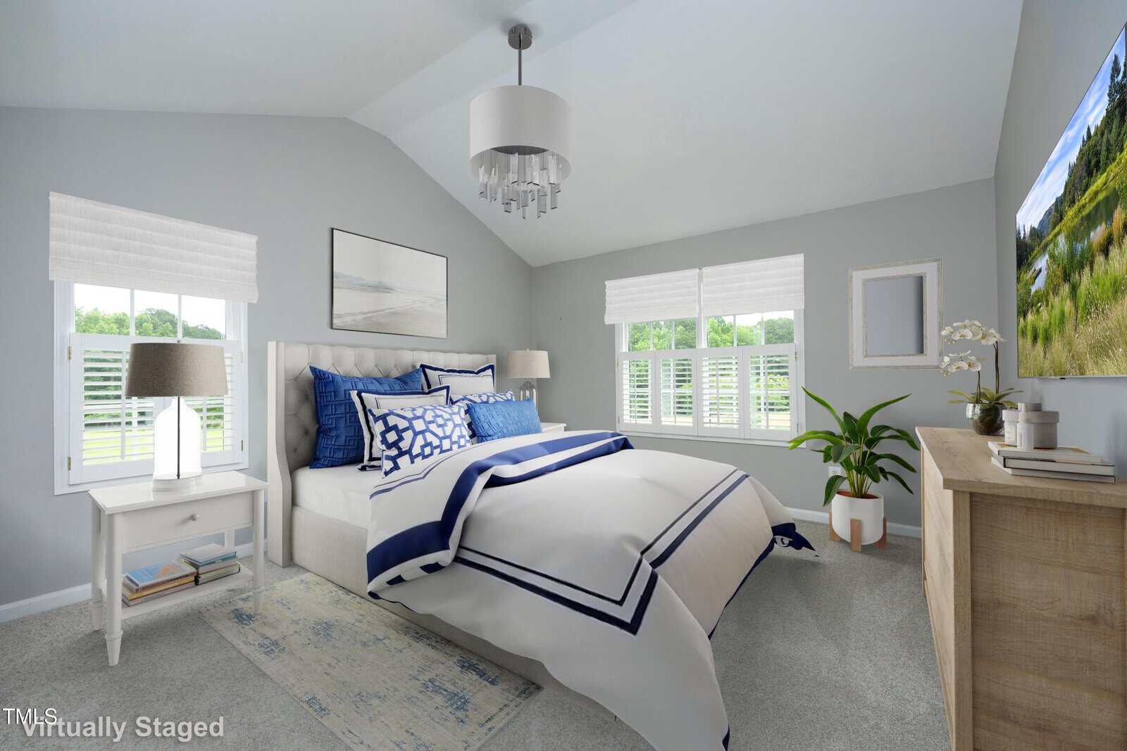 A bright bedroom with light gray walls, a bed with blue and white bedding, two white nightstands, a wooden dresser, a wall-mounted TV, and windows with white blinds. Text in the corner reads "Virtually Staged.