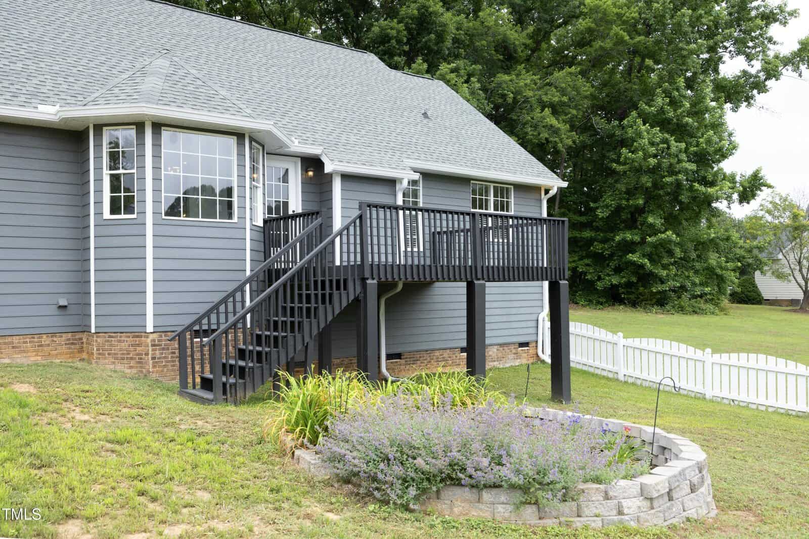 A gray house with white trim features a raised black wooden deck with stairs leading to the yard. Below the deck, there is a small circular stone flower bed with plants. A white picket fence runs along the lawn.