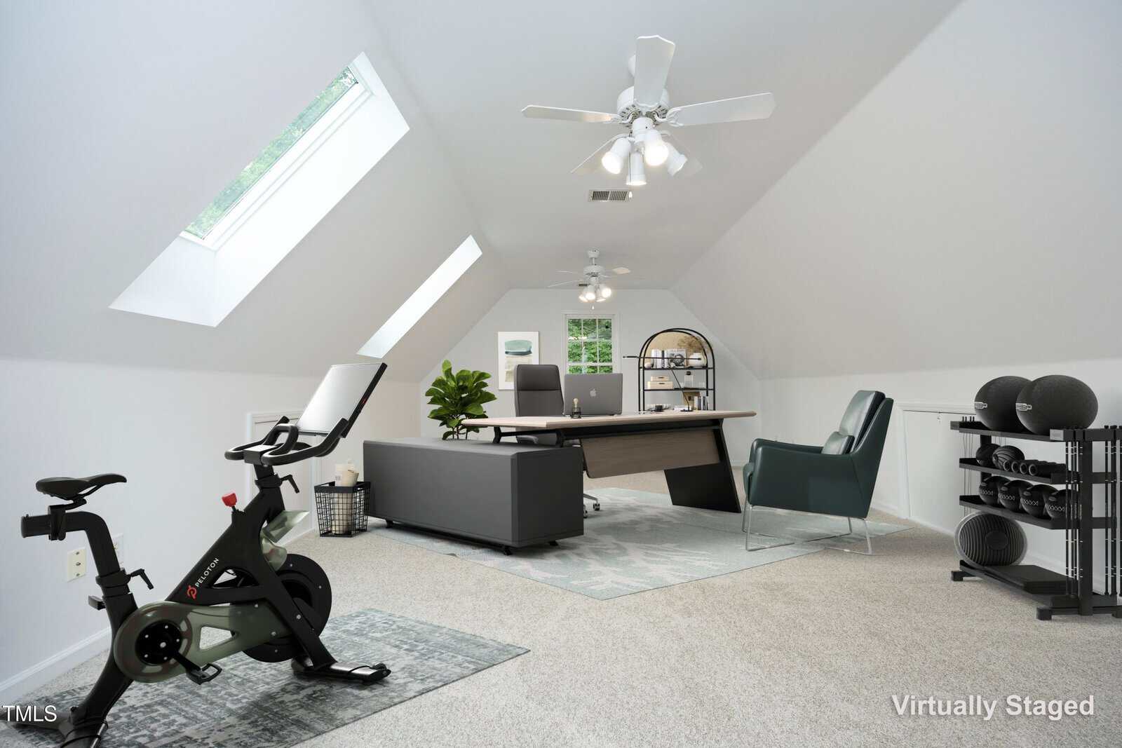 A bright attic room with sloped ceilings, a large skylight, and white walls, staged as a home office and workout area with a desk, seating, stationary bike, yoga mats, and storage racks.