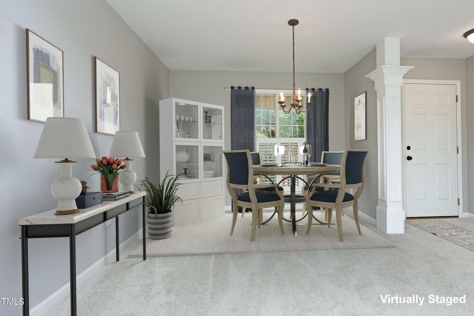 A dining room with a round table and four blue chairs on a rug, a white hutch, console table with lamps and decor, gray walls with framed art, chandelier, large window with dark curtains, and a white front door.