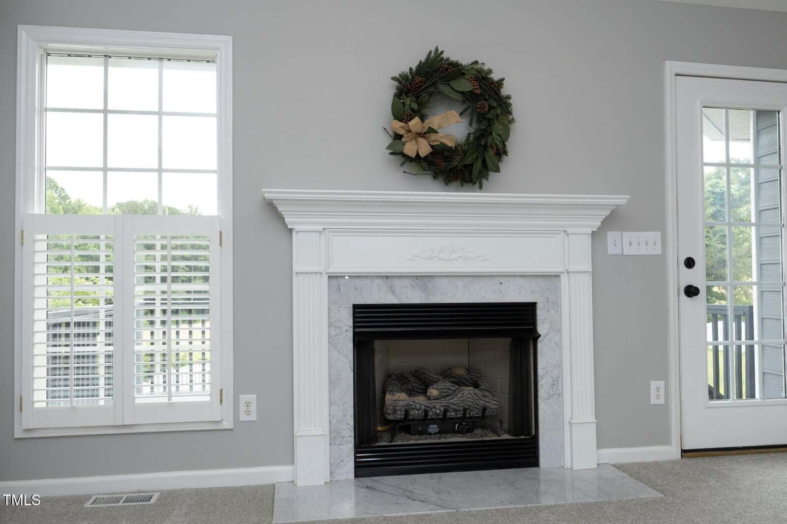 A white mantel fireplace with a marble surround is centered between a large window with shutters and a glass door. A decorative wreath hangs above the mantel on a light gray wall.
