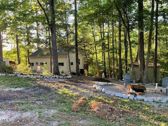 A small yellow house sits among tall trees with sunlight filtering through. In the foreground, a fire pit is surrounded by Adirondack chairs and a stone border lines a path through the wooded yard.