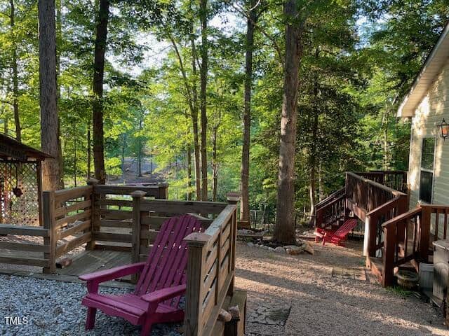 A wooden deck and stairs surrounded by trees with two red Adirondack chairs, gravel paths, and a glimpse of a wooded area in the background. Sunlight filters through the trees onto the scene.