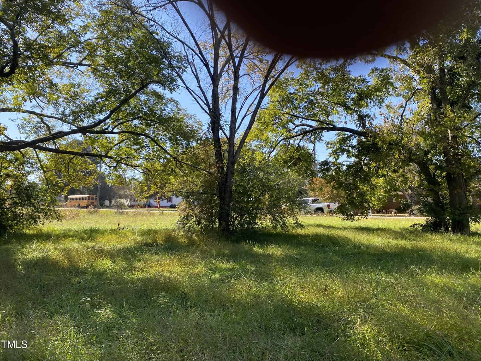 A grassy field with tall trees in the foreground, a yellow school bus, white vehicle, and house in the background. Part of the image is obscured by a finger in the upper right corner.