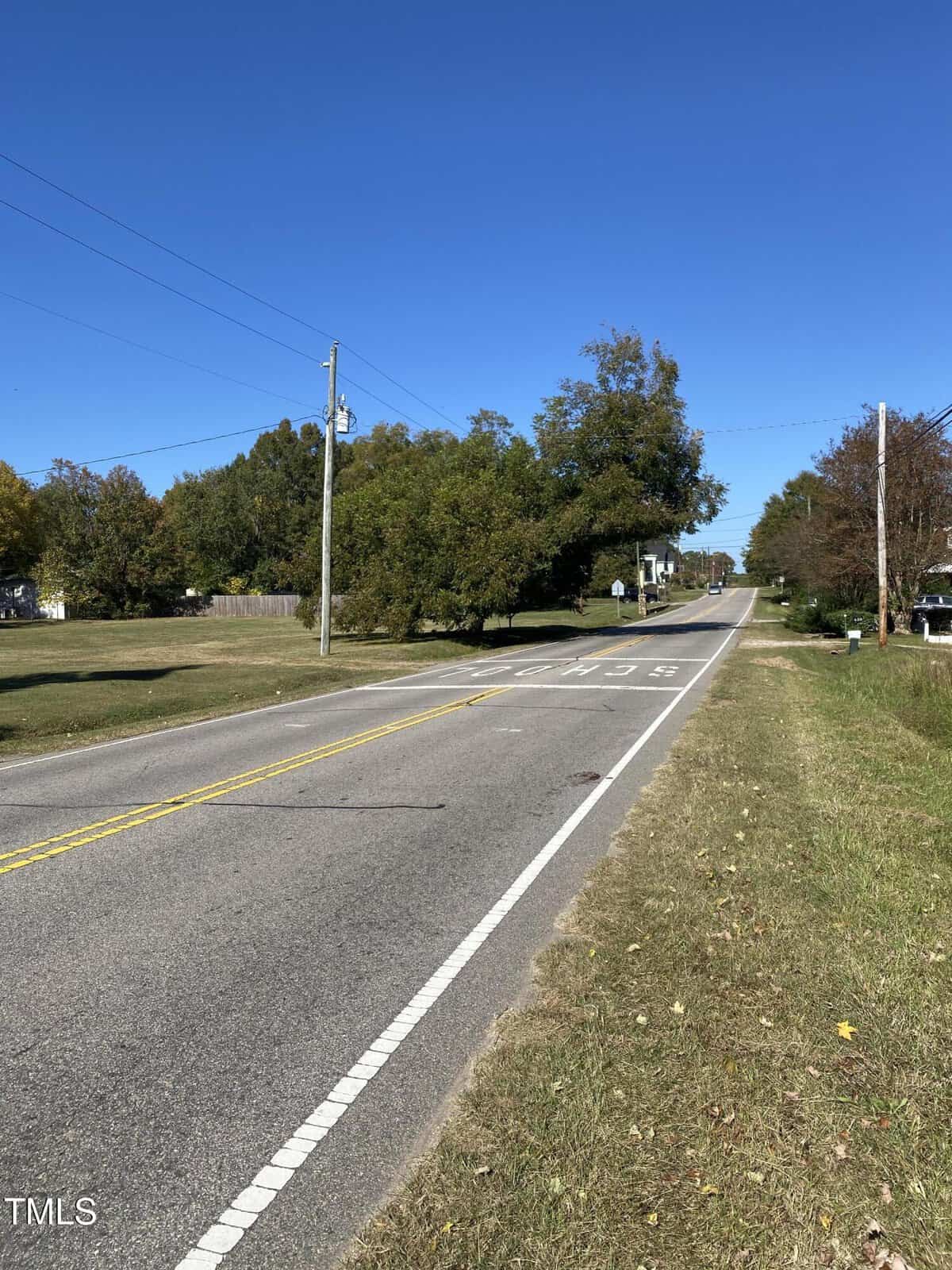A two-lane road stretches into the distance under a clear blue sky, bordered by grass, trees, utility poles, and houses. Faded white road markings and a yellow center line are visible.
