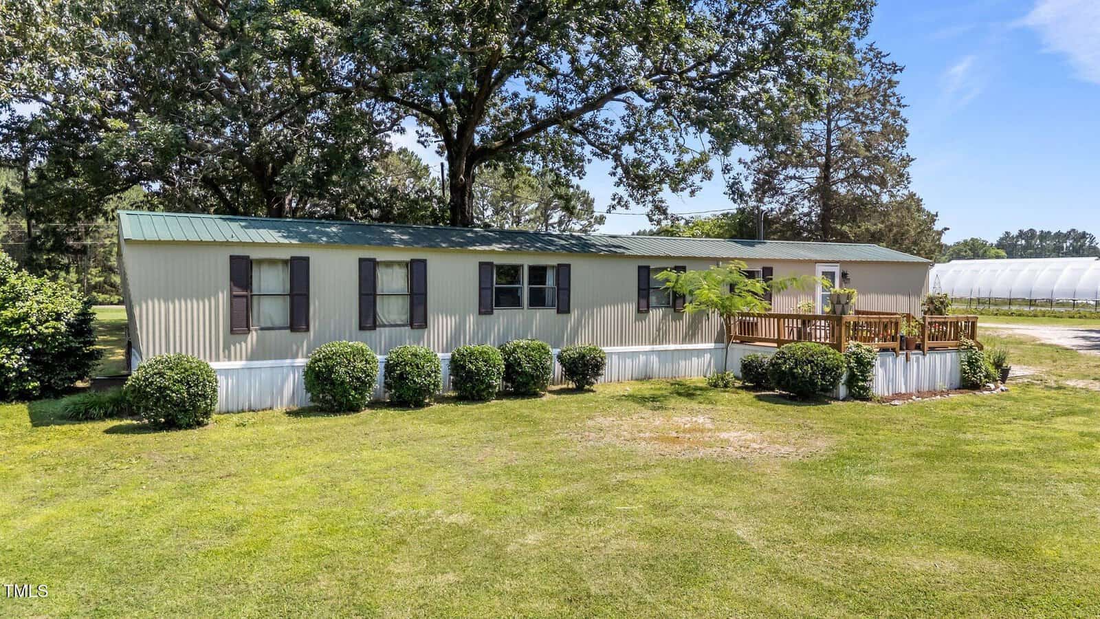 A beige mobile home with dark shutters sits on a large grassy lawn, surrounded by small bushes. A wooden porch is attached to the front, and trees and blue sky are visible in the background.