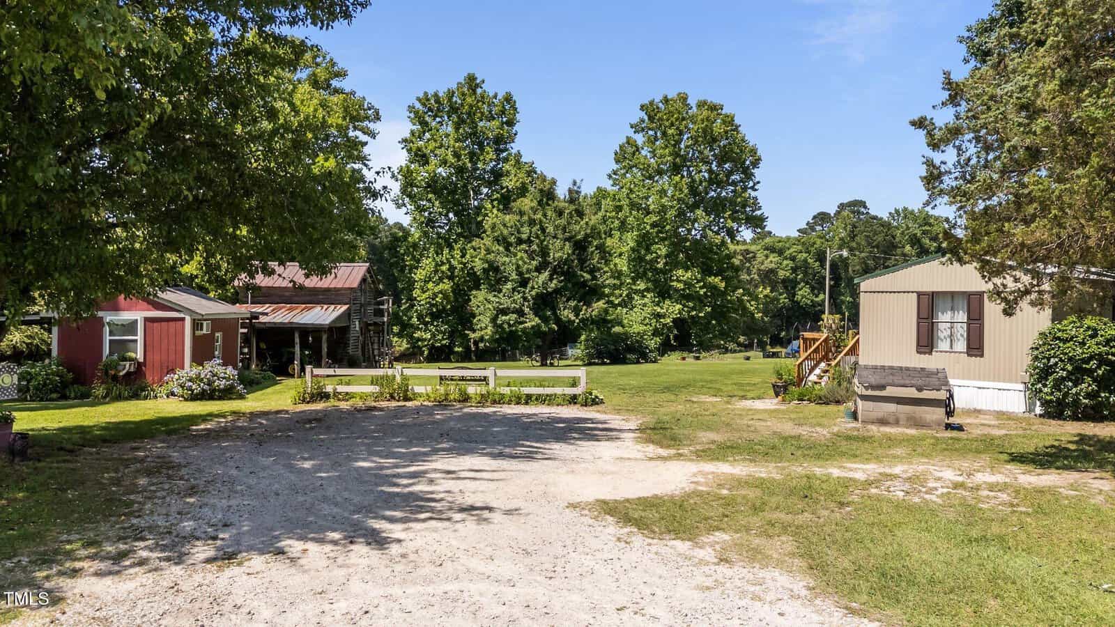 A rural yard with a gravel driveway, a red barn, a wooden shed, and a beige mobile home surrounded by green grass, trees, and a clear blue sky.