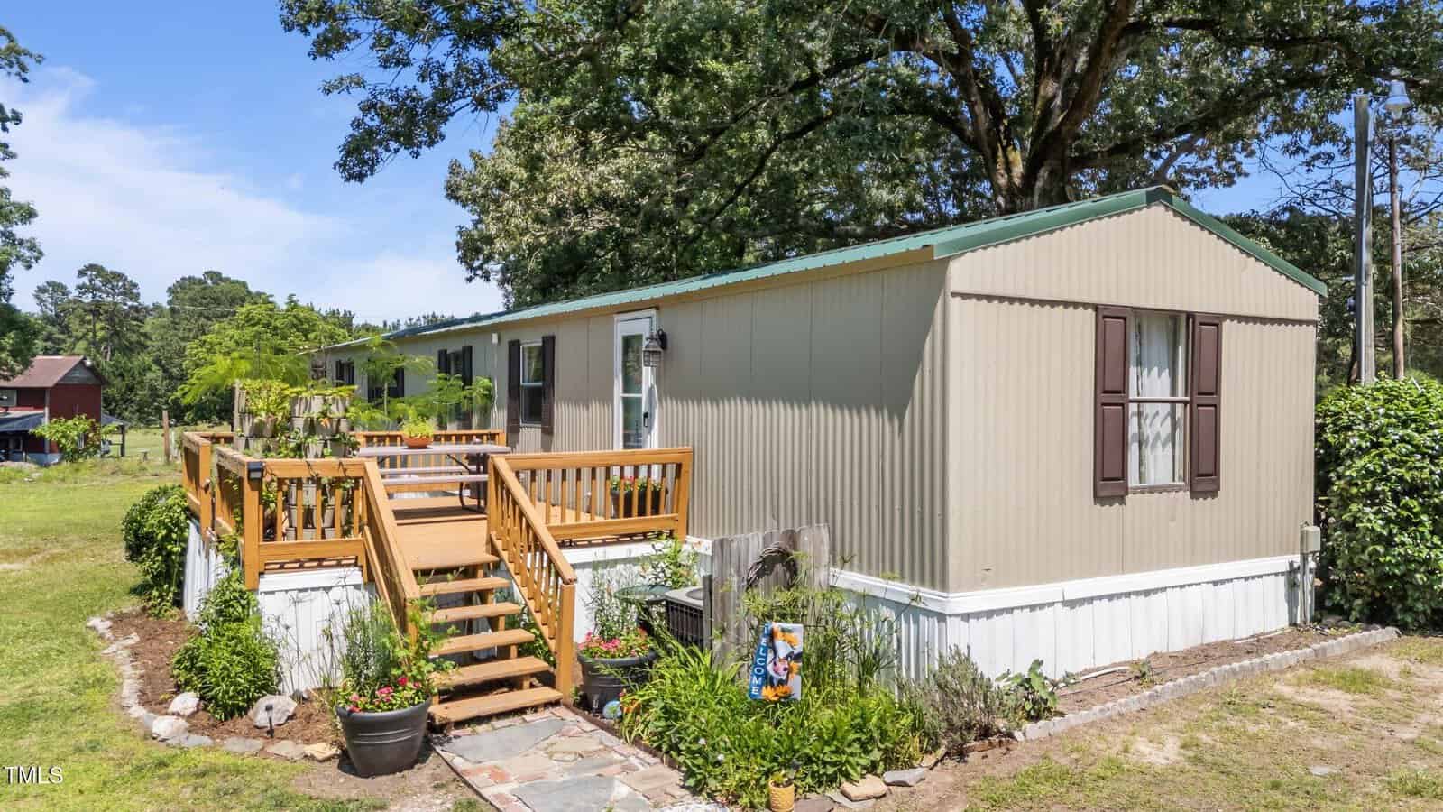 A beige mobile home with brown shutters sits on a grassy lot. A wooden deck with railings and potted plants is attached. Trees, shrubbery, and a barn are visible in the background under a blue sky.