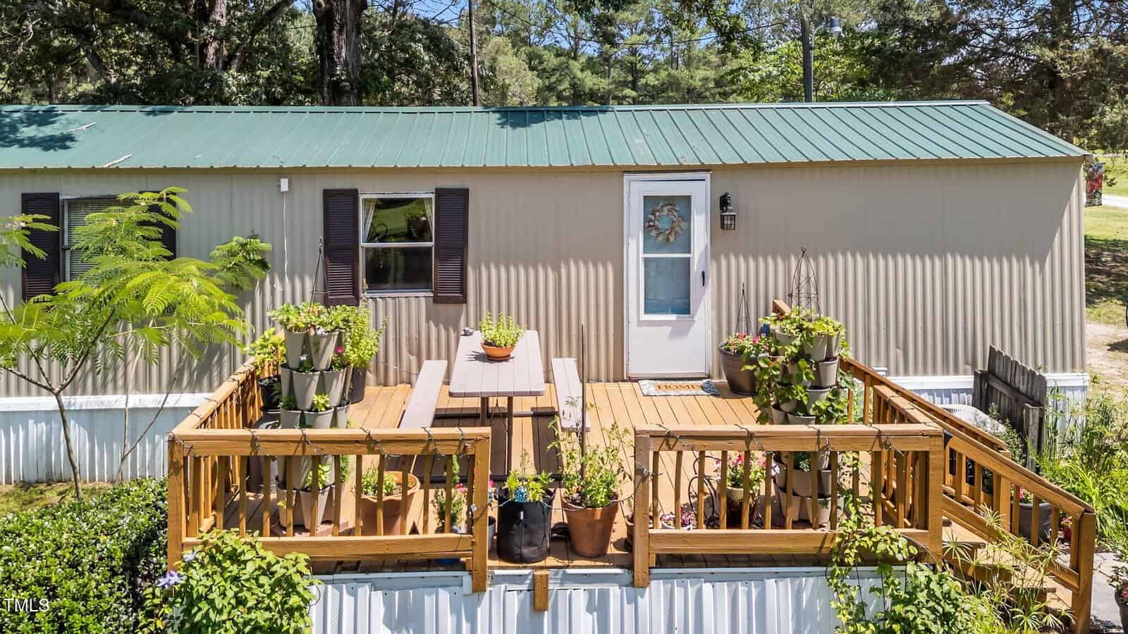 A beige mobile home with a green metal roof features a wooden deck with railings, potted plants, a picnic table, and a door with a decorative wreath. Trees and greenery surround the area.