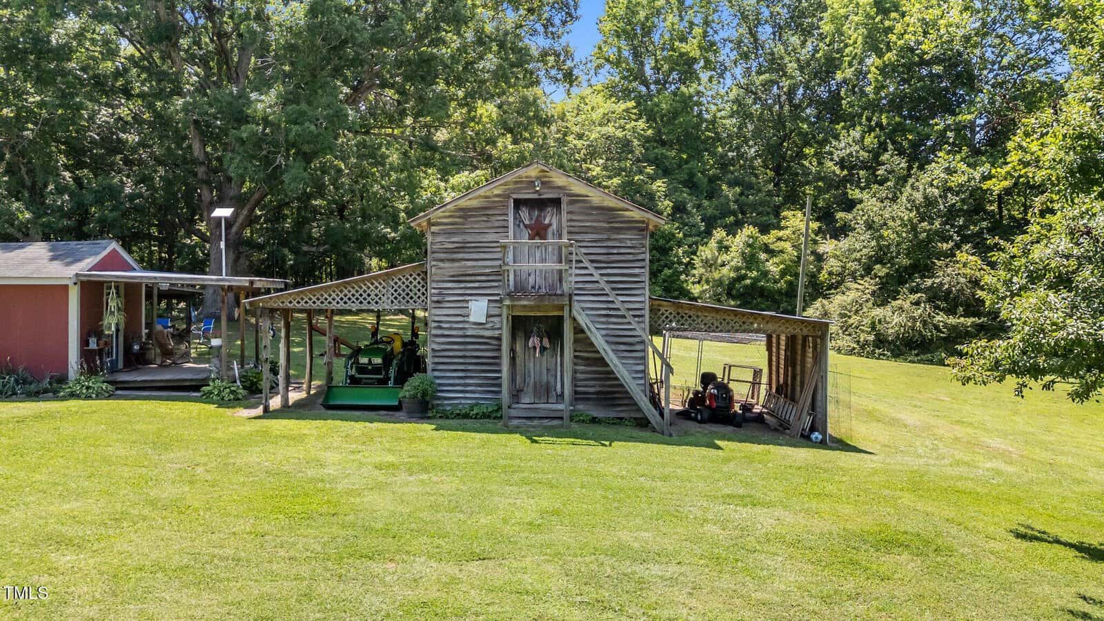 A wooden barn with a front staircase and attached side shelters sits on a grassy lawn, surrounded by trees. Lawn equipment is stored under the shelters. A small red shed is partially visible on the left.