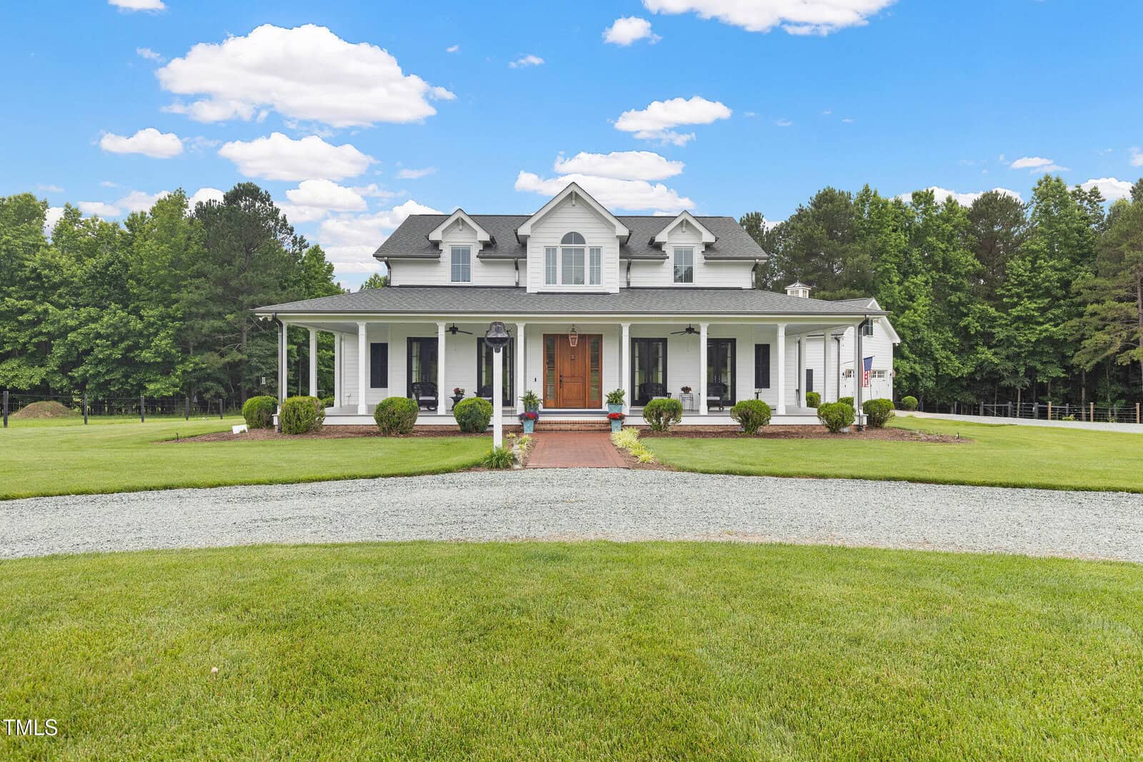A two-story white house at 4737 Uzzle Road, Rougemont, with a covered front porch, dormer windows, and a brick walkway leading to the wooden front door. Lush grass, shrubs, and trees surround the home under a blue sky with clouds.