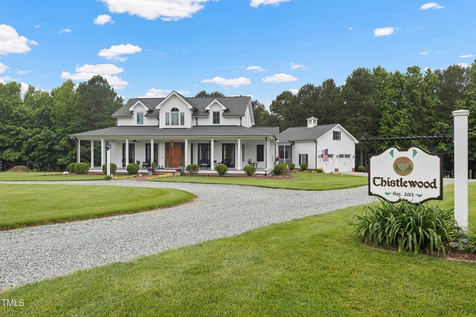 A large white house with a porch, surrounded by green lawn and trees, sits behind a gravel driveway. In the foreground, a sign reads “Thistlewood, Est. 2013.” Blue sky with scattered clouds overhead.