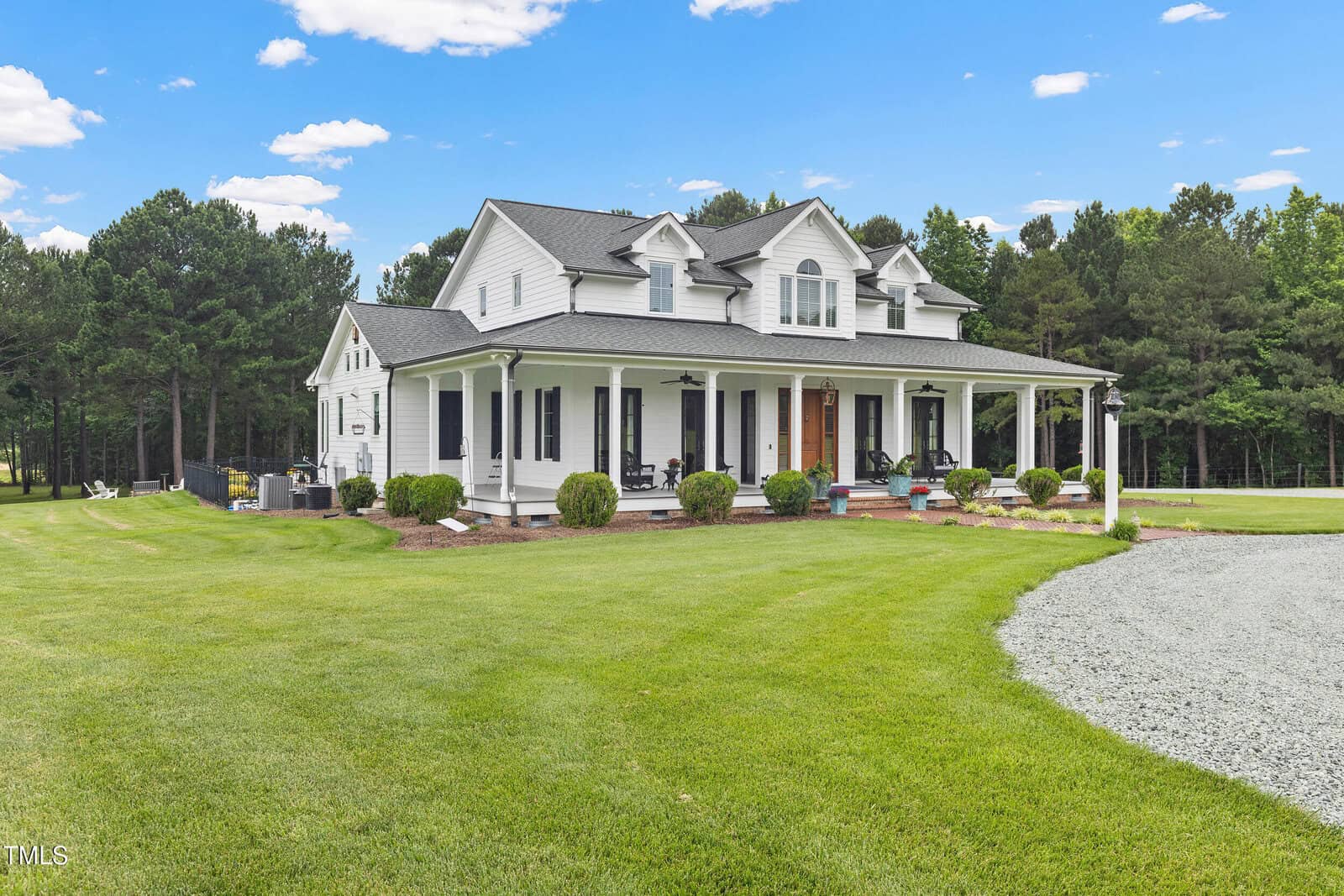 A white two-story house with a wraparound porch sits on a large, well-manicured lawn, bordered by trees. The house has multiple windows, dormers, and a gravel driveway leading to the front steps.