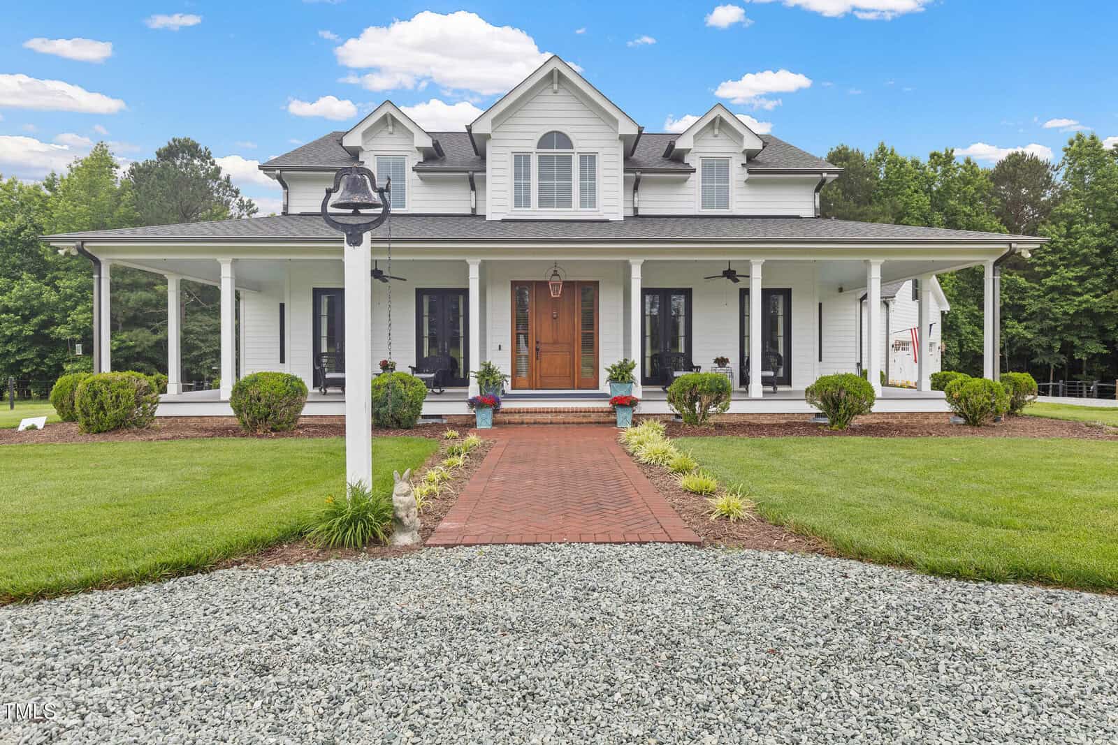 A white two-story house with a large covered porch, brick walkway, and wooden front door sits amid a well-manicured lawn, shrubs, and trees under a blue sky with scattered clouds.