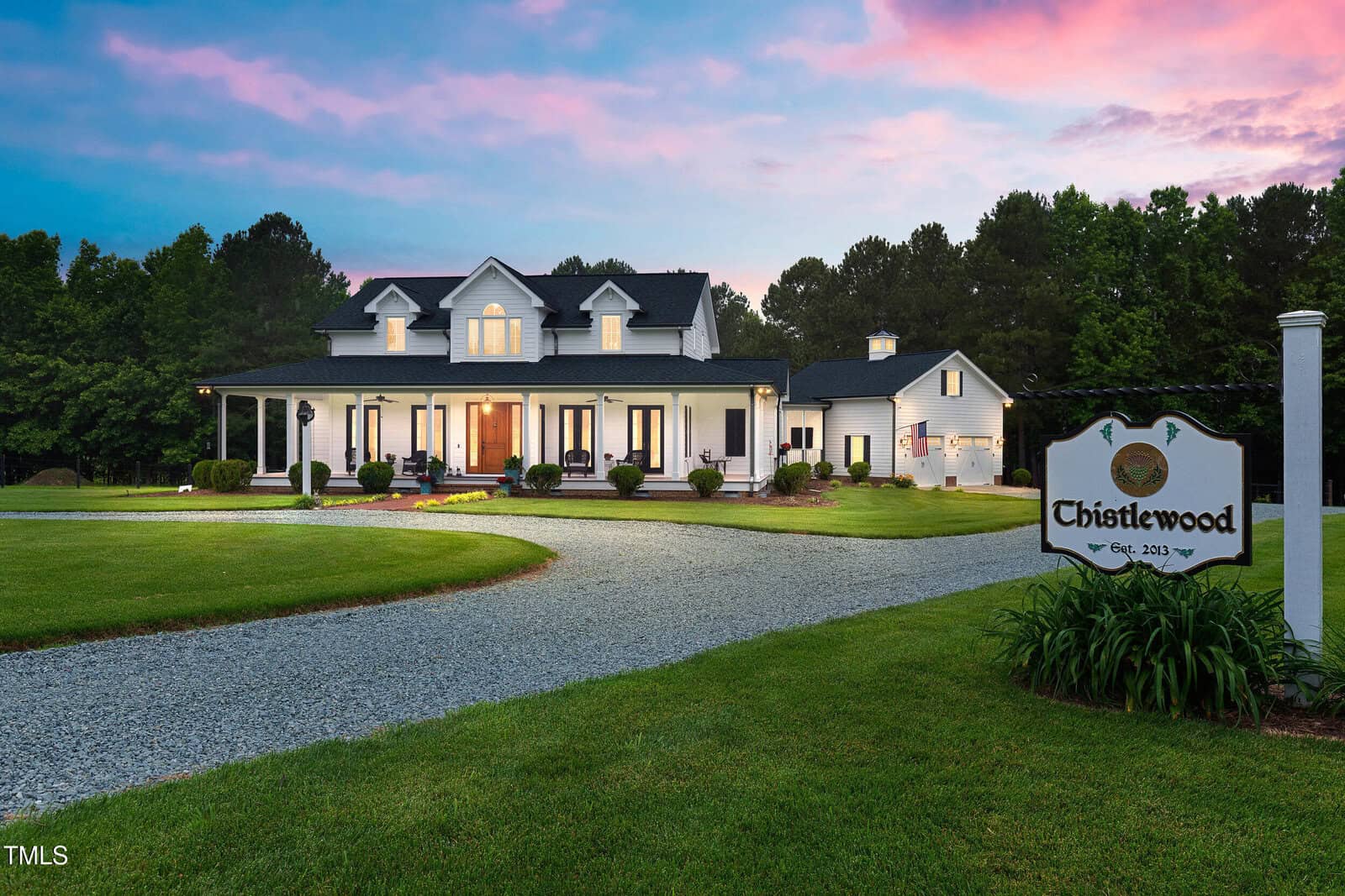 A large white house with a porch, surrounded by green lawn and trees at sunset. A curved gravel driveway leads to a sign reading “Châtelwood, Est. 2019.”.