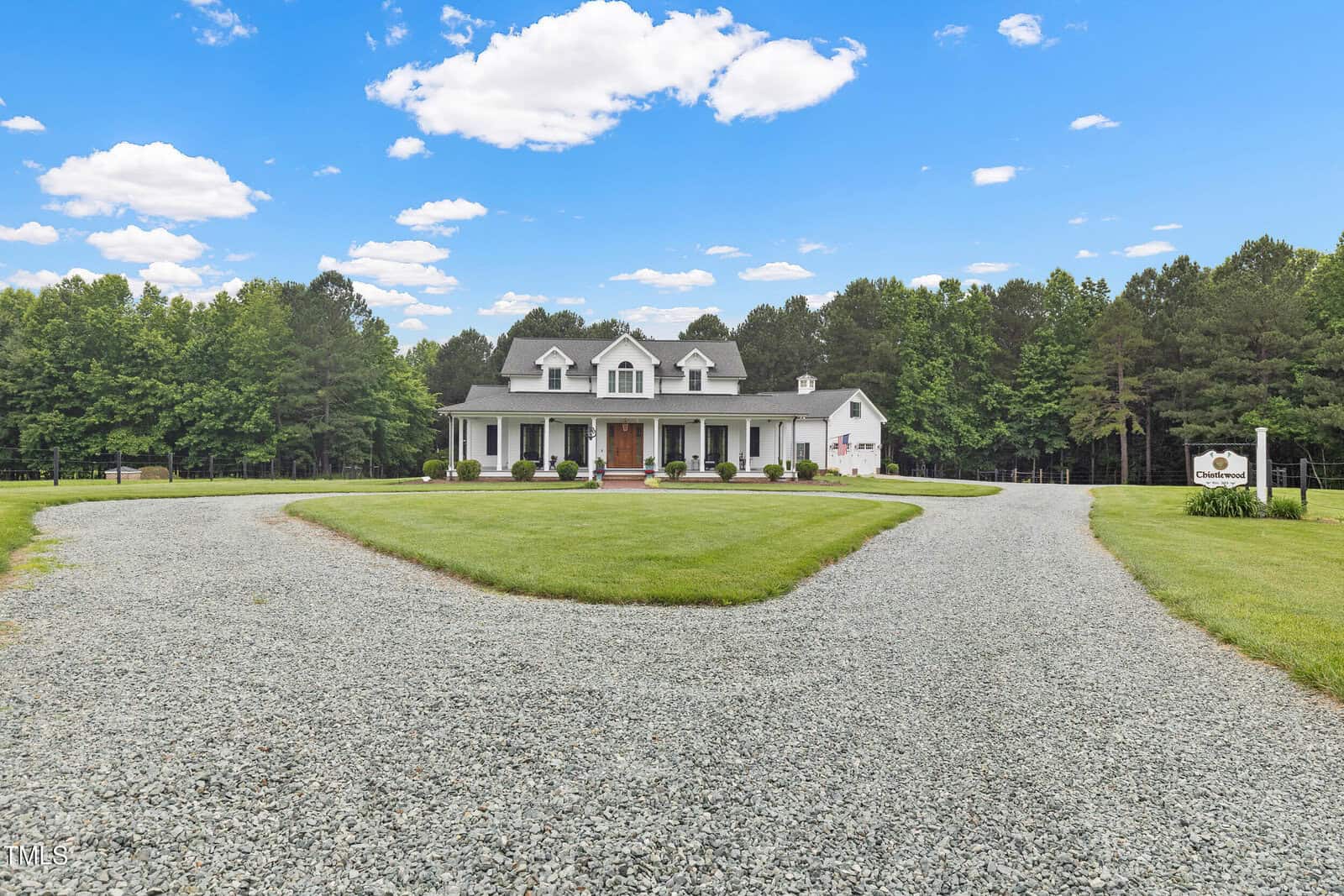 A white, two-story house with a covered front porch sits at the end of a circular gravel driveway, surrounded by a green lawn, trees, and a blue sky with scattered clouds.