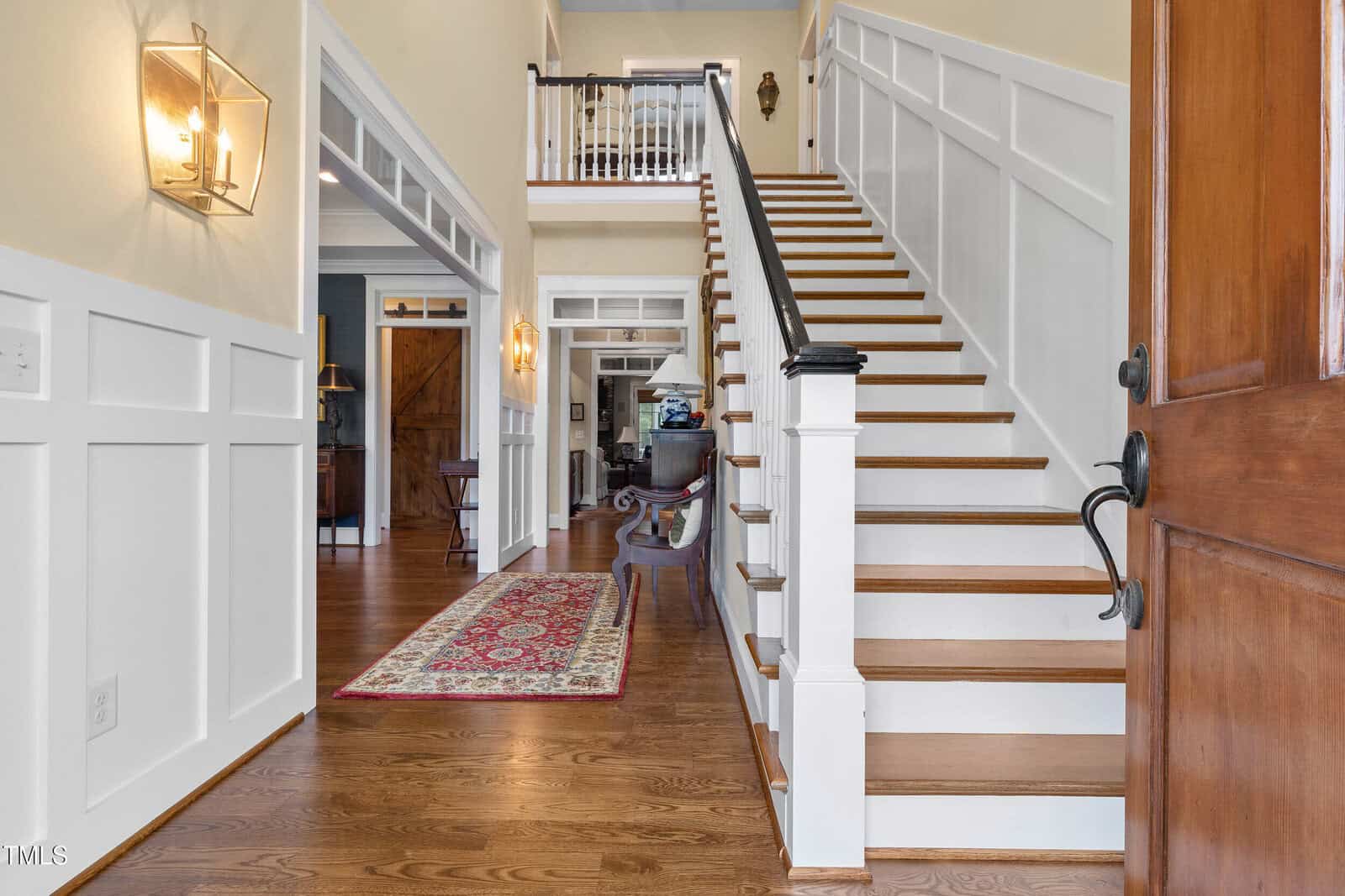 Open wooden front door reveals entry hallway with wood floors, stairs with white risers and dark bannister, wall paneling, a red patterned rug, and a glimpse into other rooms with furniture in the background.