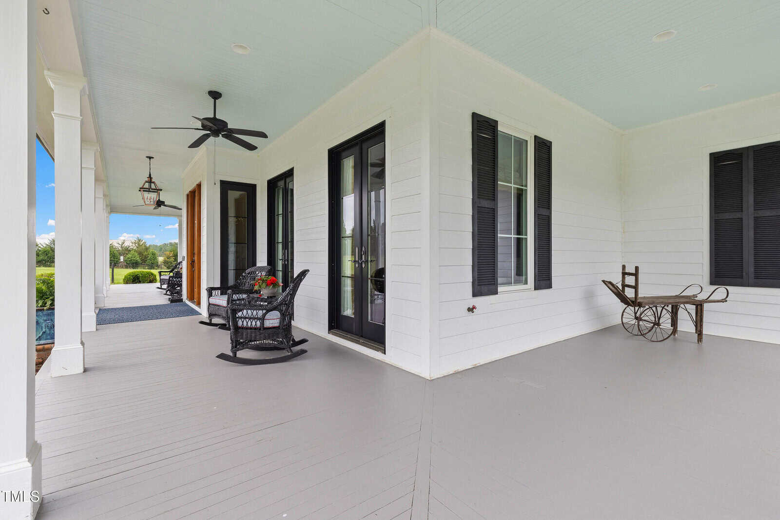 A spacious covered porch with white siding, black shutters, a ceiling fan, rocking chairs, a small table with a plant, and a decorative vintage wheelbarrow. The flooring is light gray wood.